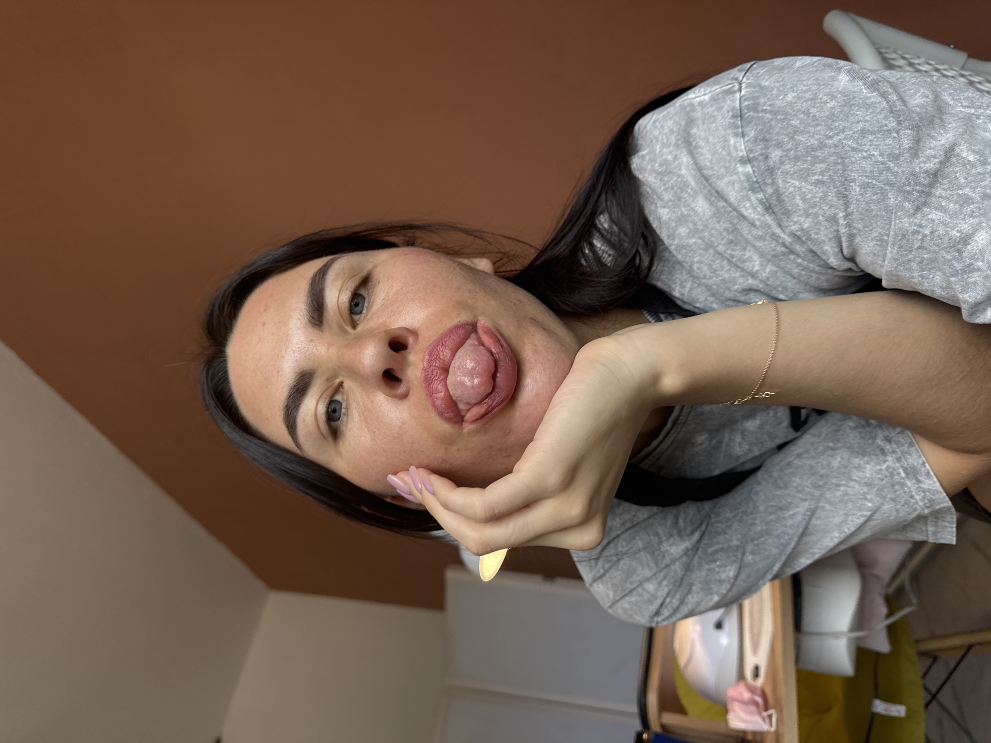 A young woman with dark hair and blue eyes is shown from the chest up, posing for a selfie in what appears to be a domestic setting. She is wearing a grey t-shirt and her left hand is propping up her chin, with her tongue playfully sticking out. Her nails are painted a light pink color. In the background, a desk is visible with what looks like a nail lamp and some pink face masks. The wall behind her is a warm, terracotta color, and part of a white ceiling is visible. The overall impression is of a casual, personal moment captured in a home environment. There is no indication of the city of Chaikovsky, Russia, in the image.