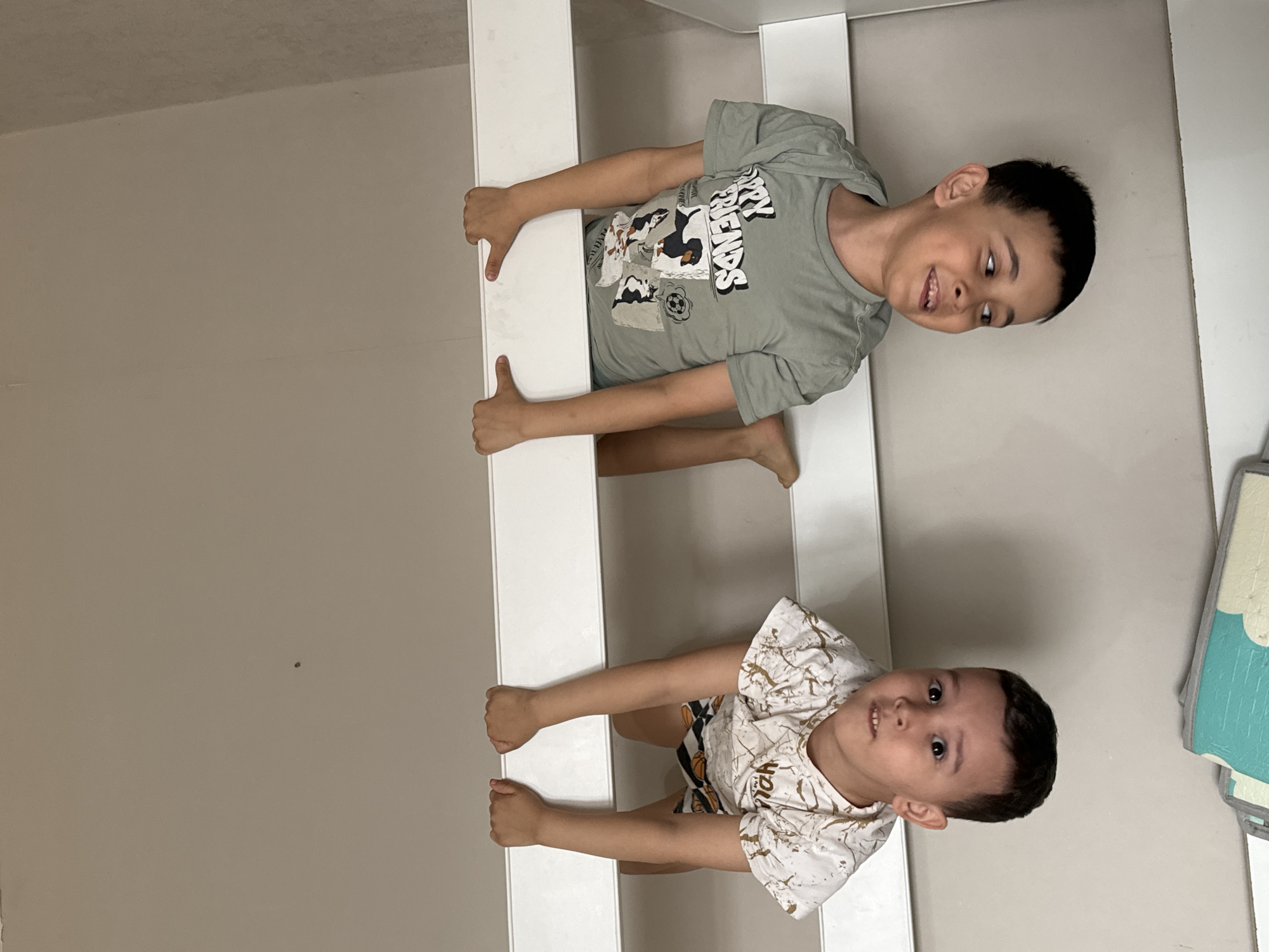 Two young boys are hanging from a white structure, possibly a bunk bed or a climbing frame. The boy at the top is wearing a light green t-shirt with a cartoon dog graphic, and the boy below has a white t-shirt with a beige marble pattern. They are both smiling and appear to be enjoying themselves. The location is in a room with light beige walls. There is a blue and white play mat visible on the right side of the image. The exact location in the city of Chaikovsky, Russia, cannot be determined from the given image.