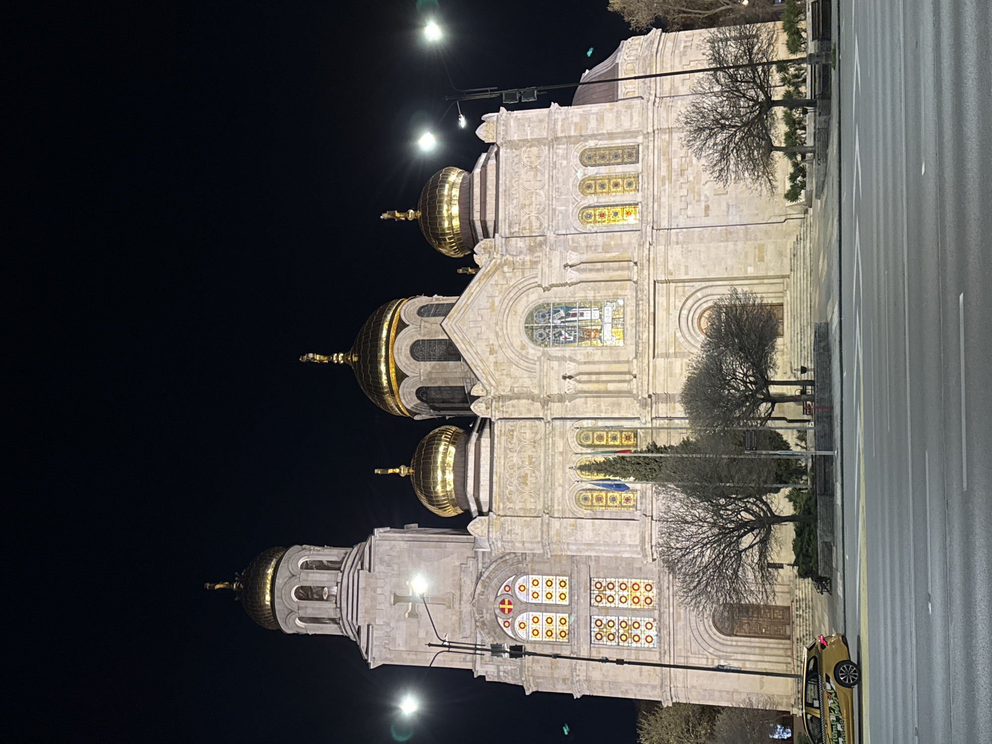 The image depicts the Dormition of the Mother of God Cathedral in Varna, Bulgaria, at night. The cathedral is illuminated and features a large facade with four prominent golden domes. The architecture is ornate with detailed stonework and stained glass windows. A street runs parallel to the cathedral, and a single taxi is parked along the curb. Several bare trees stand in front of the building. The background is dark, with streetlights illuminating the area.