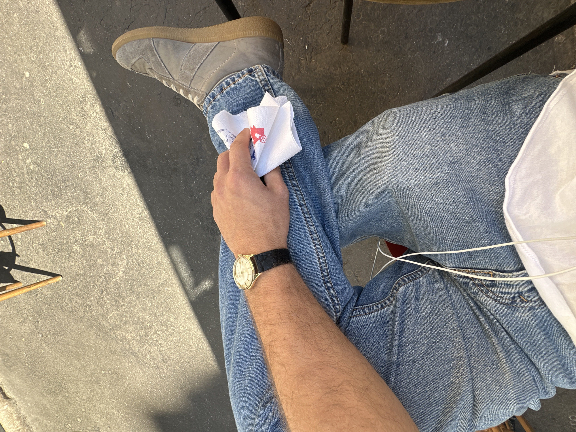 A first-person, overhead perspective image depicts a person seated outdoors, wearing light blue jeans, a grey suede sneaker, and a gold wristwatch with a black leather strap. The individual holds a folded white paper napkin in their left hand. A white t-shirt is visible on the right. The setting appears to be an outdoor cafe or similar establishment, judging from the presence of chair legs and a dark, paved surface. The location cannot be definitively confirmed as Milan, Italy based on the provided image alone.