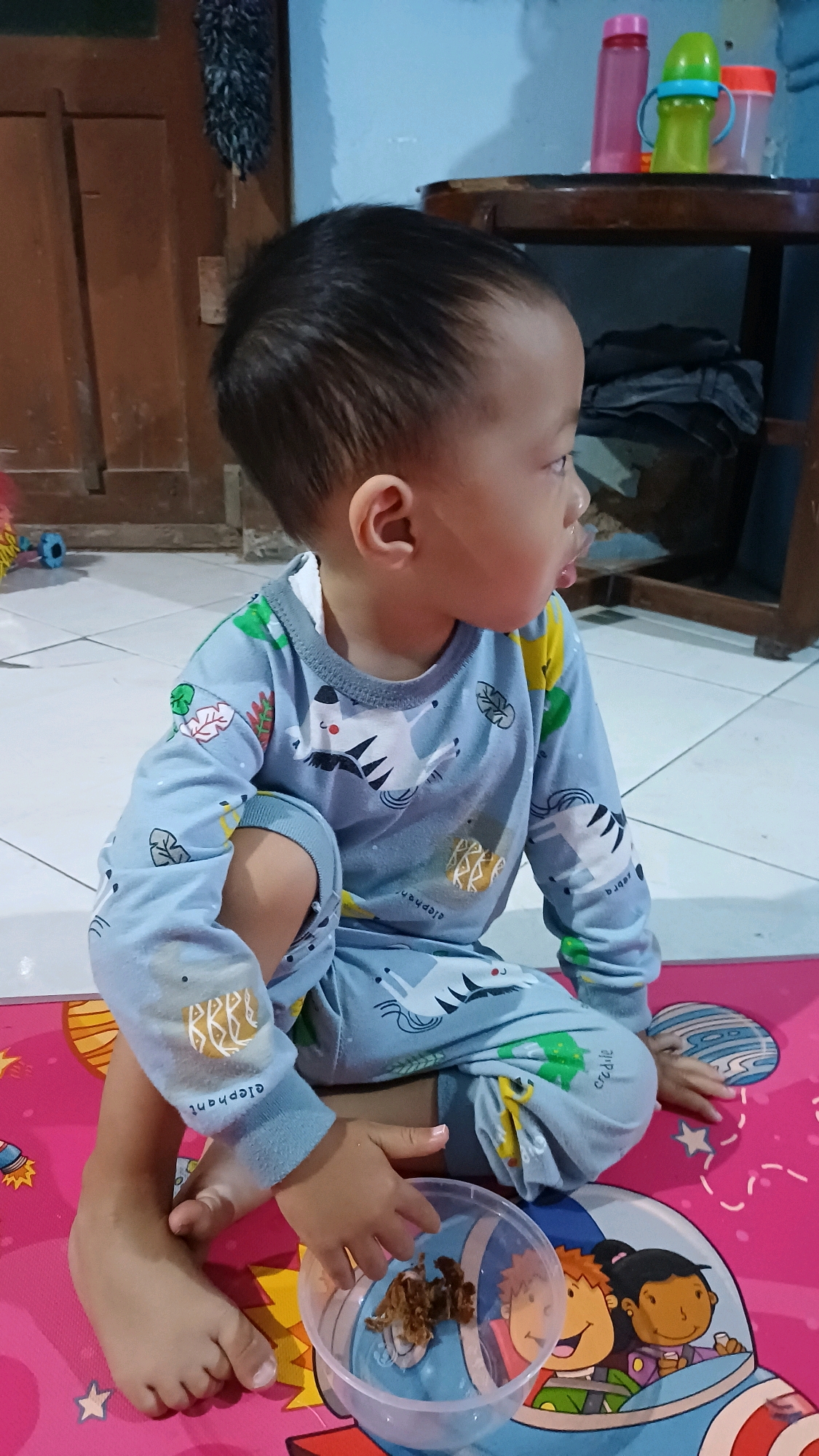 A young child, likely under the age of three, is sitting on a pink mat with cartoon rocket ship graphics. The child is wearing light blue pajamas with a repeating pattern of zebras and elephants. The child is looking to the right, with their tongue slightly protruding. In front of the child is a clear plastic bowl containing a small amount of brown, granular food. The child's right hand is reaching into the bowl. The child's left foot is visible, resting on the mat. The background shows a tiled floor, a wooden door on the left, and a table with several colorful bottles on the right. The scene appears to be indoors, possibly in a home in Wonokerso, Indonesia.