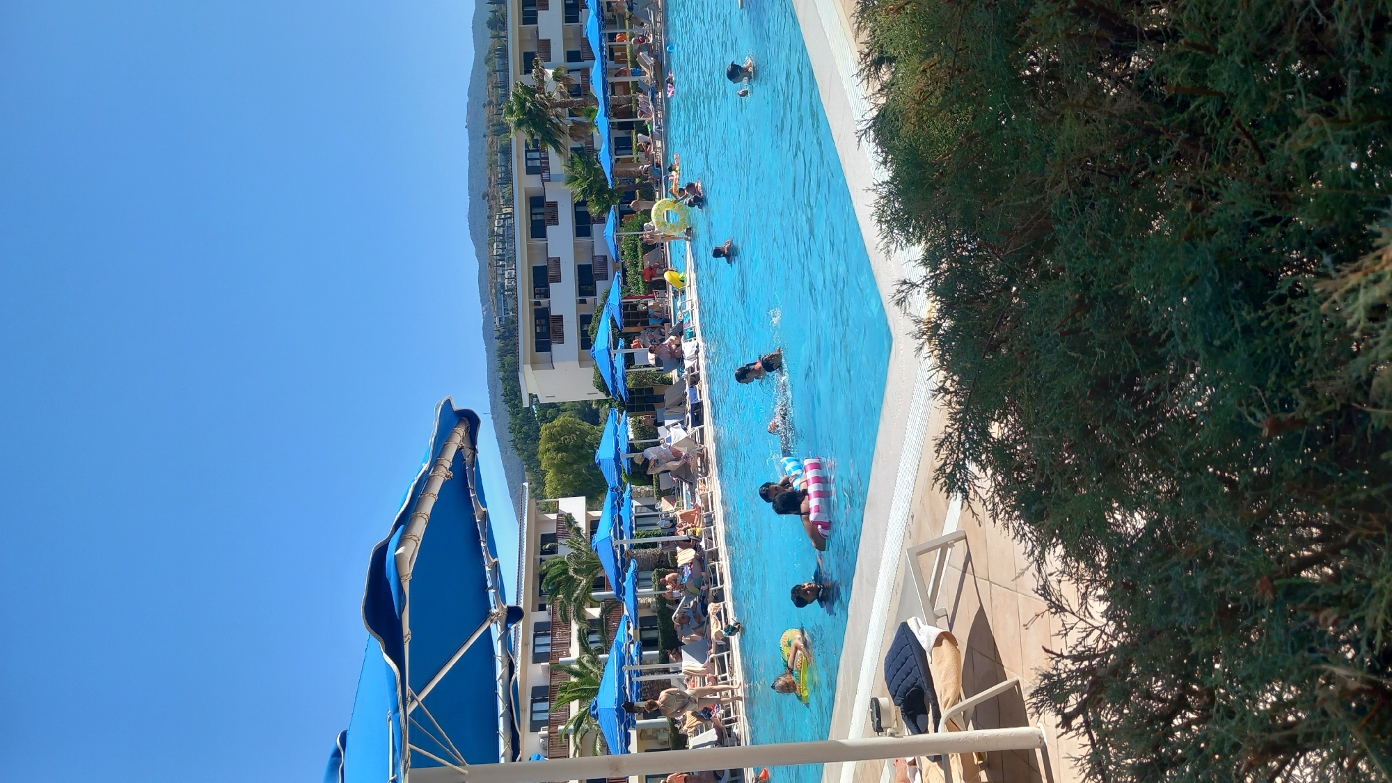 This is a daytime, eye-level, full shot of a crowded outdoor swimming pool area at a resort in Kos, Greece. The pool is filled with clear blue water, and several people are swimming or floating on inflatable rings. In the foreground, a large blue umbrella shades part of the pool area. In the background, there are resort buildings, palm trees, and a hillside with some vegetation. The sky is clear and blue. There are lounge chairs and umbrellas scattered around the pool, with many people relaxing on them.