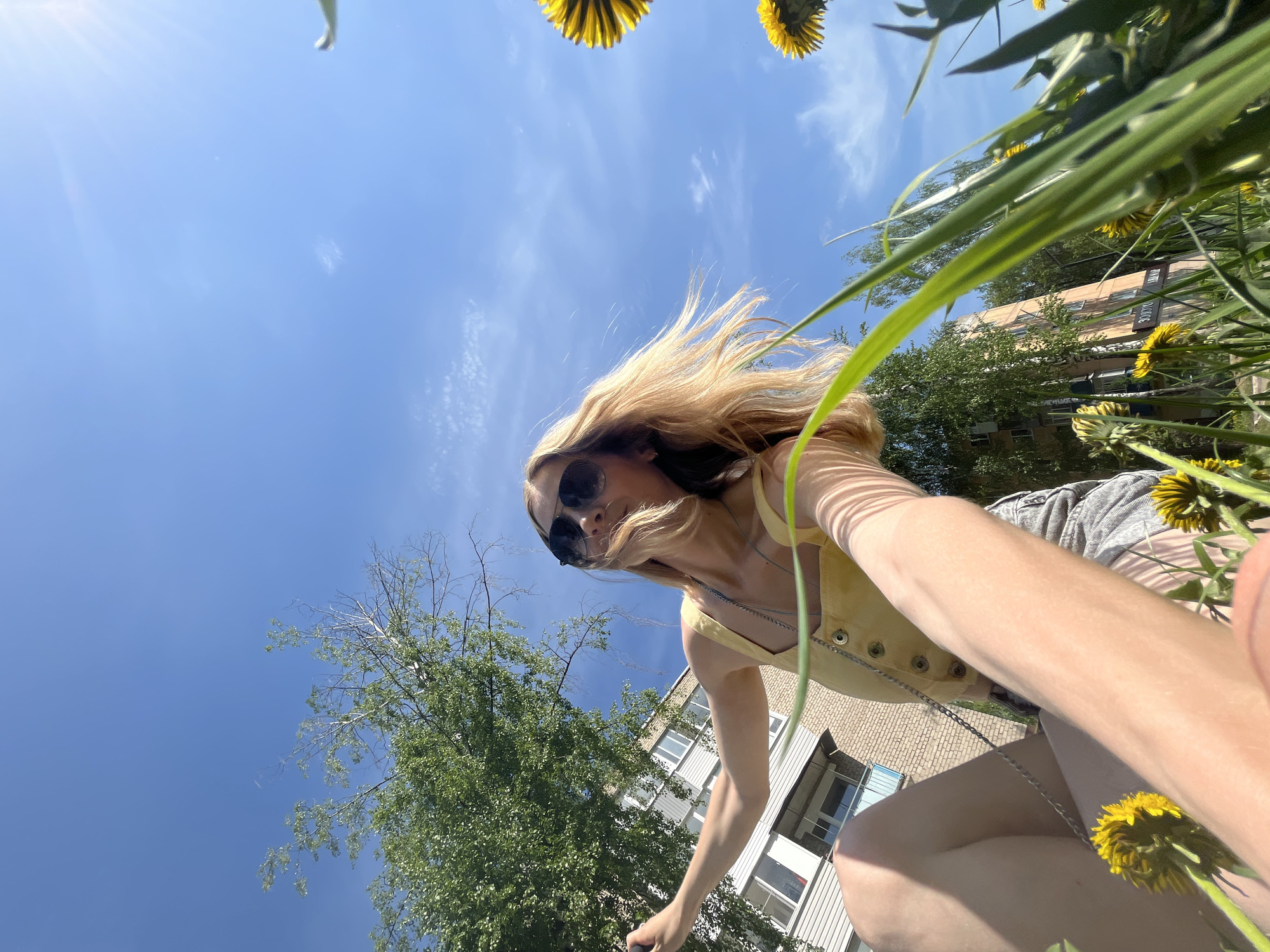 A low-angle, upward-facing photograph shows a woman, seemingly taking a selfie, against a background of a sunny blue sky and buildings. She is wearing sunglasses and a yellow top with a silver chain. Her blonde hair is partially obscuring her face. The presence of grass and yellow flowers frames the image, suggesting an outdoor location. The background includes a brick building with white-framed windows and a green tree. Considering the city is Chaikovsky, Russia, the buildings appear to be residential. There are no visible actions or interactions apart from the act of taking a selfie.