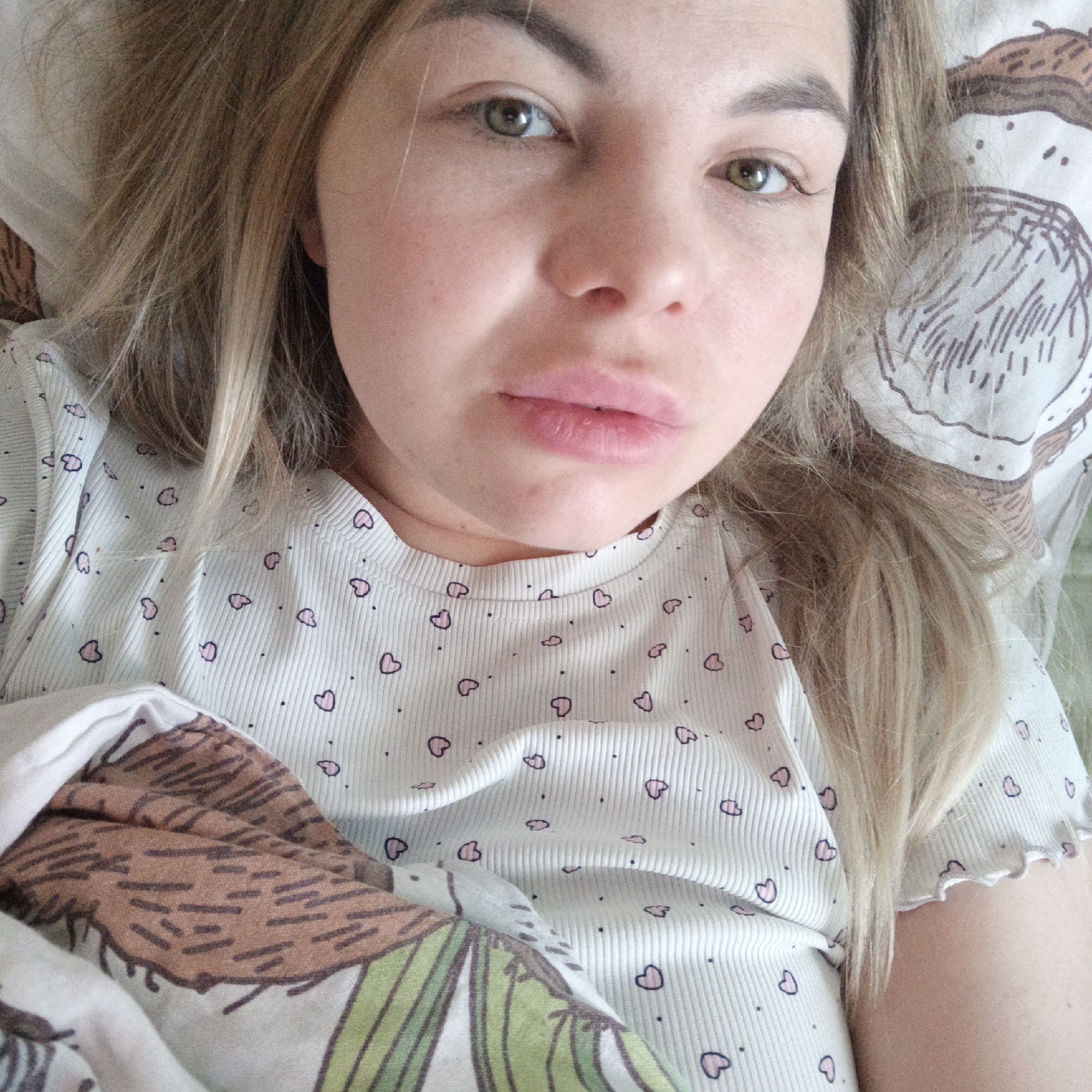 A woman lies in bed, wearing a white shirt with small pink hearts. She is partially covered by a comforter with a pattern of coconuts and leaves. The location is Unknown, Unknown.