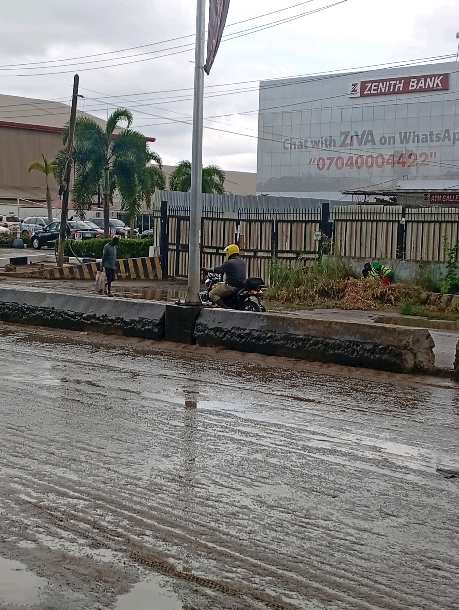 The image depicts a street scene in Ikeja, Nigeria. In the foreground, a wet, muddy road shows tire tracks and puddles, indicating recent rainfall or poor drainage. A concrete barrier separates the road from the sidewalk. On the sidewalk, a person wearing a green shirt and carrying a white bag walks away from the camera, while another person wearing a yellow helmet rides a motorcycle and is partially visible. Behind them, a building with the "ZENITH BANK" logo is prominent, featuring text that reads "Chat with ZiVA on WhatsApp" and a phone number. A black metal fence separates the bank building from the street. Palm trees are visible behind the fence, and the sky is overcast. Two individuals in the background appear to be engaged in some form of manual labor or tidying overgrown vegetation near the fence. A car is parked to the left of the scene.