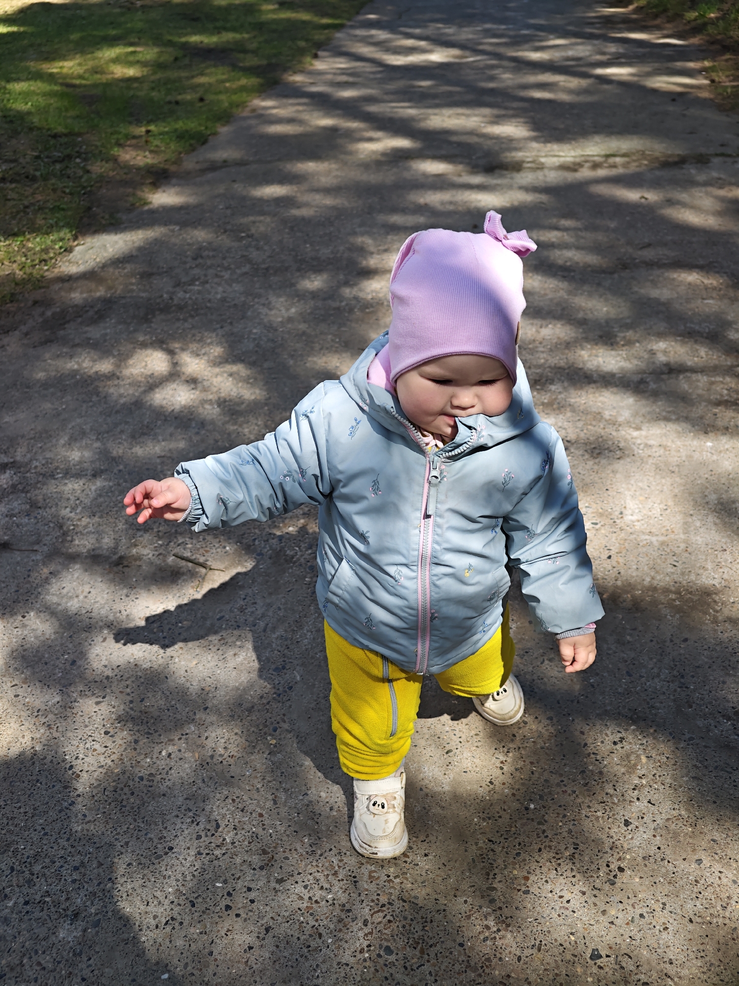 A toddler wearing a pink hat with a bow, a light blue jacket with a pink zipper and yellow pants is walking on a concrete path. The child is wearing white shoes with a cartoon face on the sides. The path has shadows from trees. The photograph was taken in the city of Chaikovsky, Russia.