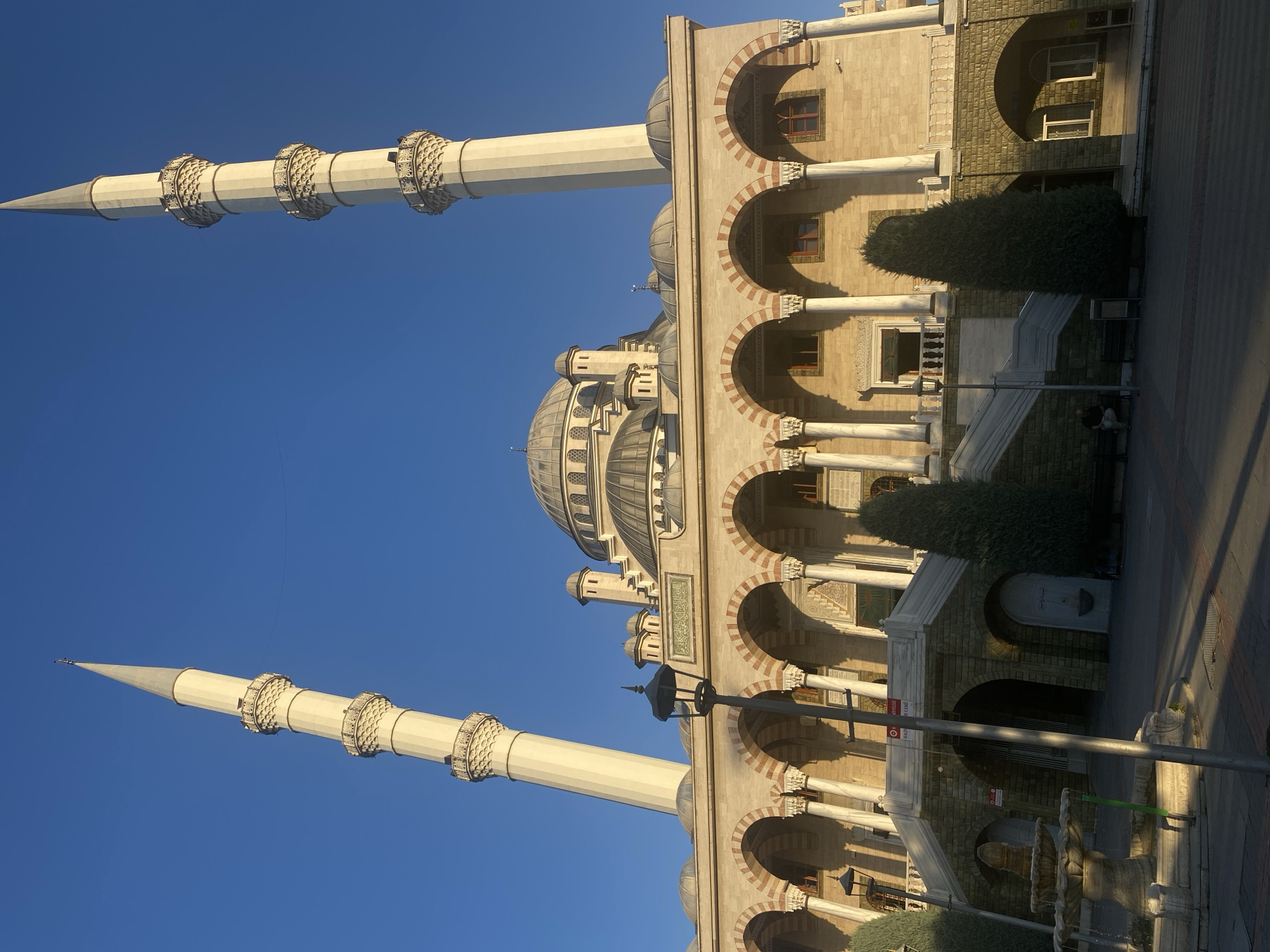 The image shows the exterior of a large mosque in Konya, Turkey, under a clear blue sky. The mosque features a central dome, multiple smaller domes, and two tall minarets with ornate balconies. The facade of the mosque is constructed from light-colored stone and is adorned with a series of arched windows and colonnades. White marble columns support the arches, and red and white patterned inlays are visible within the arches. A grand set of stairs leads up to the main entrance, flanked by cypress trees. To the left of the stairs, a stone fountain with multiple tiers is visible. In the foreground, a lamppost stands on the paved ground, and a few people are seated on benches. The lighting suggests it is either early morning or late afternoon, with long shadows cast across the ground. The mosque appears to be a significant architectural landmark within the city.