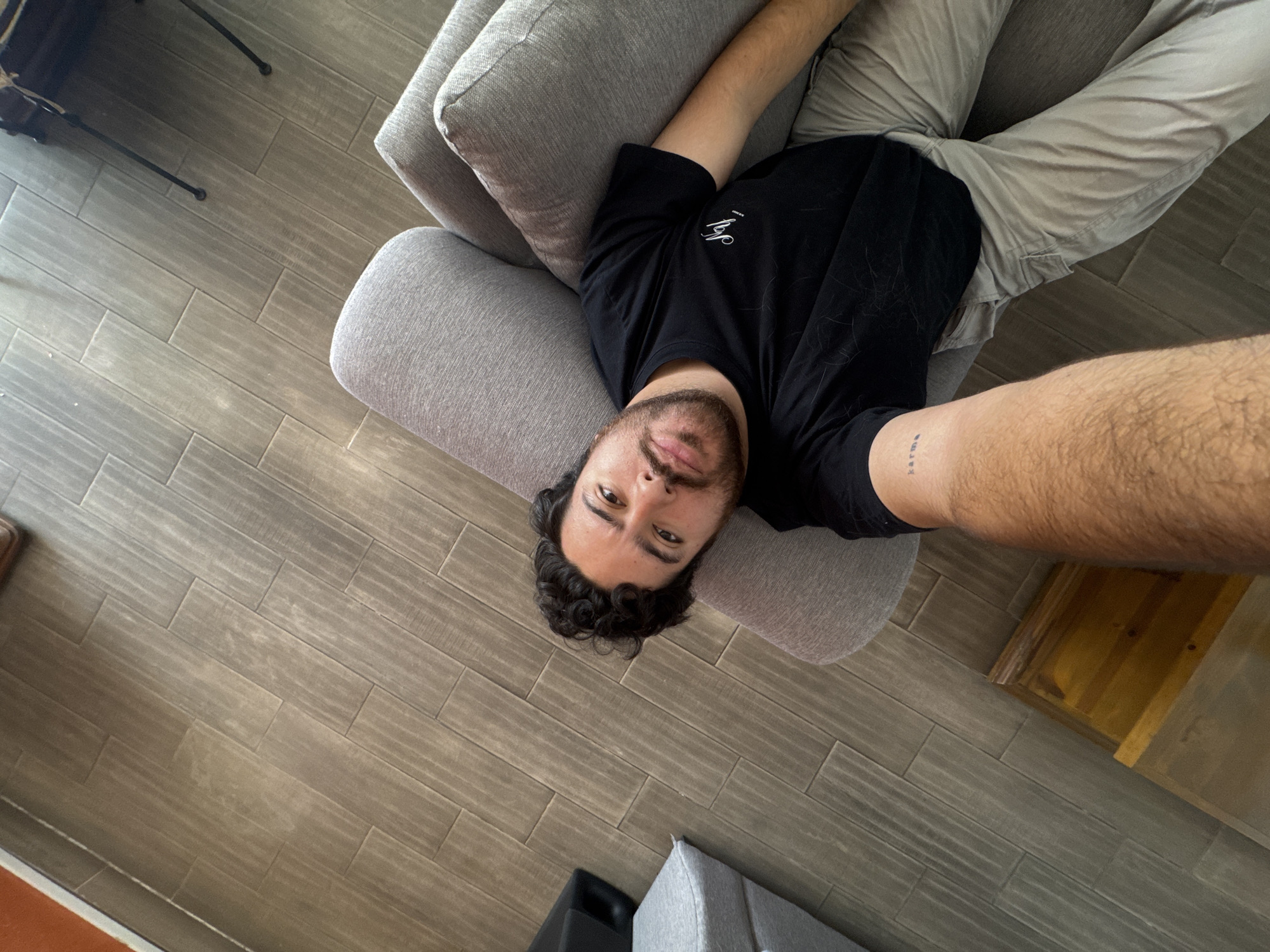 A top-down shot shows a person lying on a gray couch. The person is wearing a black t-shirt and light-colored pants. The background is a tiled floor. The tattoo "Karma" is visible on the person's right forearm. Based on the style of the floor tiles and furniture, and the presence of the person, it is most likely an indoor residential space in the city of Puebla, Mexico.