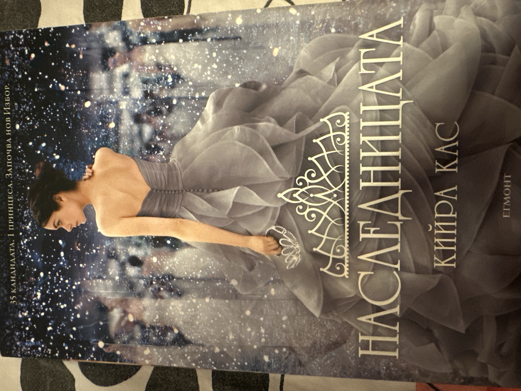 This book cover depicts a serene woman in an elegant, flowing grey strapless gown. Her dark hair is styled up, and she is looking down and to her left with a pensive expression. In her right hand, she delicately holds a sparkling tiara or crown. The background is a dark, dreamlike scene, possibly a grand ballroom or hall, with numerous small white sparkles resembling falling snow or glitter, creating a magical atmosphere. Blurred figures are visible in the distance, suggesting a social event. The woman's gown flows around her, hinting at movement.
Visible text in Bulgarian includes the title "НАСЛЕДНИЦАТА" (The Heir/Heiress), the author "КИЙРА КАС" (Kiera Cass), and a tagline on the left side: "35 КАНДИДАТА. 1 ПРИНЦЕСА. ЗАПОЧВА НОВ ИЗБОР." (35 CANDIDATES. 1 PRINCESS. A NEW SELECTION BEGINS.). The publisher's name, "ЕГМОНТ" (Egmont), is also visible at the bottom right. The overall impression is one of elegance, fantasy, and royalty, likely for a young adult romance or dystopian novel.
