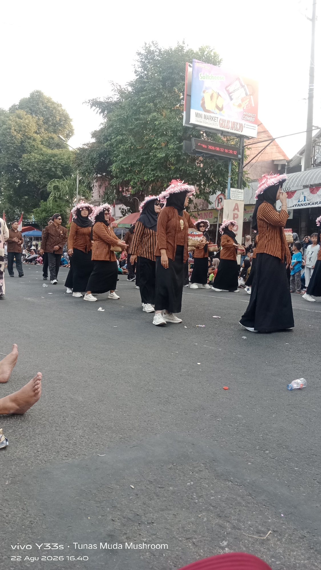 On a paved street, a group of people in traditional clothing are participating in a parade. The individuals are wearing brown and black outfits with decorative hats. The background shows trees, buildings, and a billboard advertising "Salicheese" and "MINI MARKET GELIS MULYO." A digital clock on a pole displays "2026/08/22 16:35:12". On the ground, there are scattered pieces of trash, including a plastic bottle and bottle caps. The image appears to be taken from a low angle, with some feet visible in the foreground. The timestamp at the bottom of the image indicates "22 Agu 2026 16.40", suggesting the event occurred on August 22, 2026, at 4:40 PM.