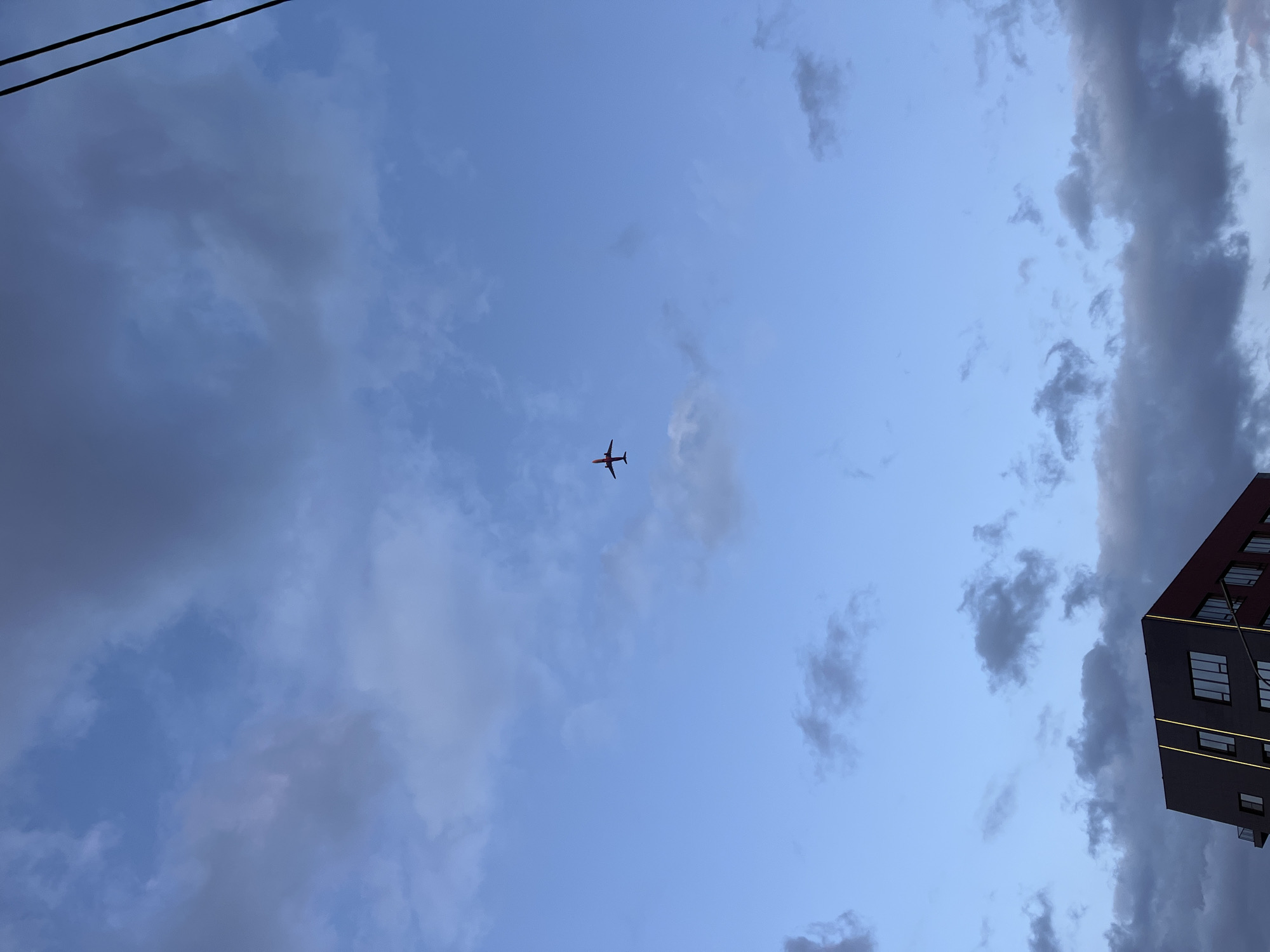 The image shows a view looking up at a blue sky with scattered clouds. An airplane is visible in the center of the frame. In the upper left corner, two power lines can be seen. The corner of a tall building is visible in the lower right side of the image. The building has a dark exterior with light-colored window frames and yellow-colored lines. The image has been taken during daytime. The location is unknown and there is no information regarding its presence in Krasnoyarsk, Russia.