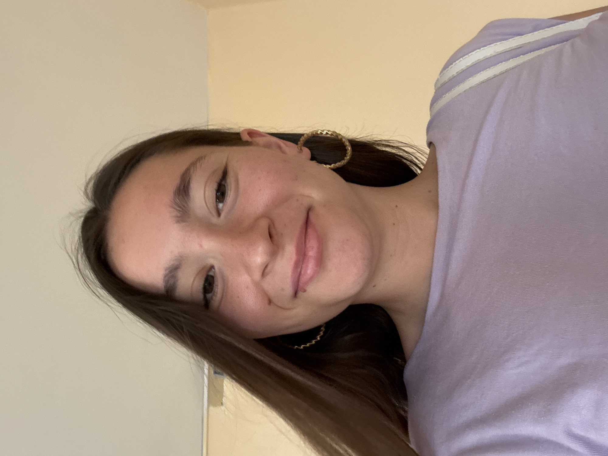 A young woman with long brown hair and freckles smiles at the camera. She is wearing a light purple t-shirt and large gold hoop earrings. Her hair is partially covering her face on the left side. The background is a light beige wall. The image appears to be taken indoors, possibly in an apartment or a room. There is no indication of Sofia, Bulgaria in the image.
