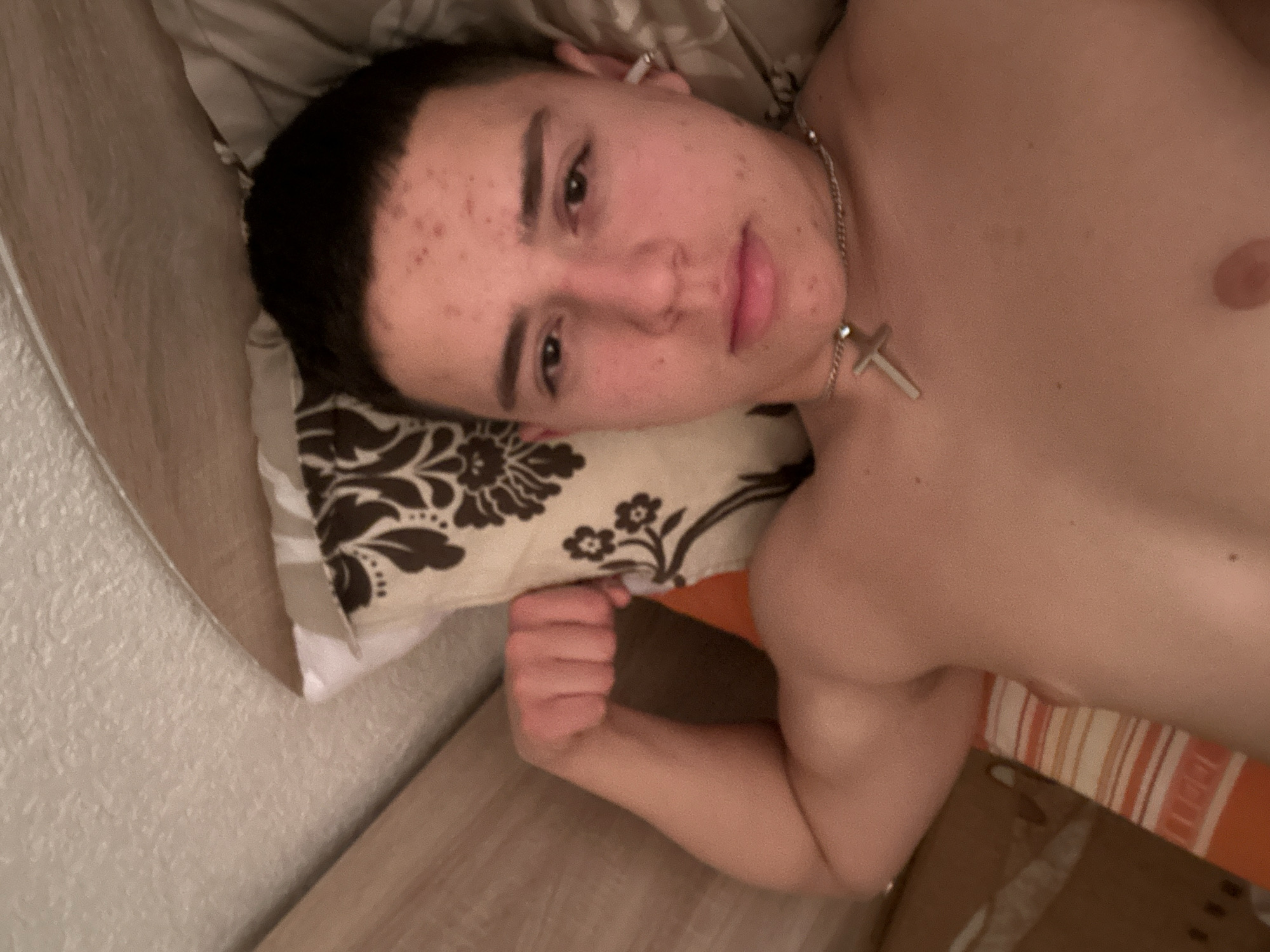 A young man lies on a bed in an indoor setting. He has short dark hair and acne on his face. He is wearing a silver necklace with a cross pendant. His arm is flexed, with his fist clenched. The bed has patterned pillows and a striped orange and white blanket. The location is unknown.