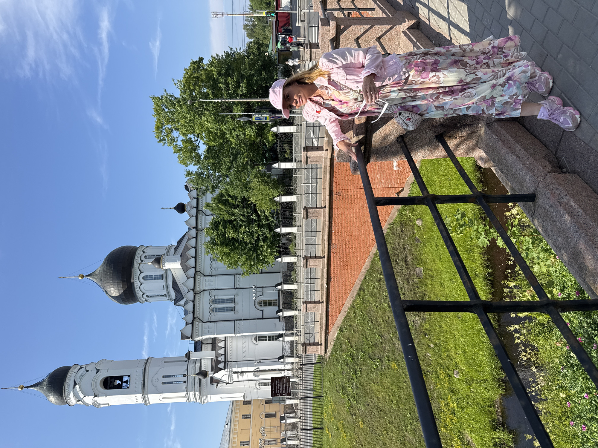 A woman is captured in a candid pose, leaning on a stone railing and looking downwards, in what appears to be a public park or plaza. She is wearing a pink cap, a pink jacket, and a long, flowing floral dress in shades of pink, white, and green. Her footwear consists of pink sneakers. A small, decorative handbag rests near her.
The scene is set outdoors, with a clear blue sky dotted with wispy clouds, suggesting a pleasant, sunny day. Behind the woman, a large, white, multi-domed church with black domes stands prominently, likely a significant landmark. The architecture of the church is ornate, featuring arched windows and intricate detailing. Adjacent to the church, lush green grass and trees add to the serene atmosphere. In the background, a road with traffic lights and people walking can be observed, indicating a connection to a more urban environment.
The overall impression is one of a bright, warm day, perhaps late morning or early afternoon, with the woman enjoying a moment of rest or contemplation amidst a historical and scenic backdrop in Lomonosov, Russia. The image is captured from a slightly elevated angle, providing a good view of both the woman and the surrounding environment. There is no visible text in the image.