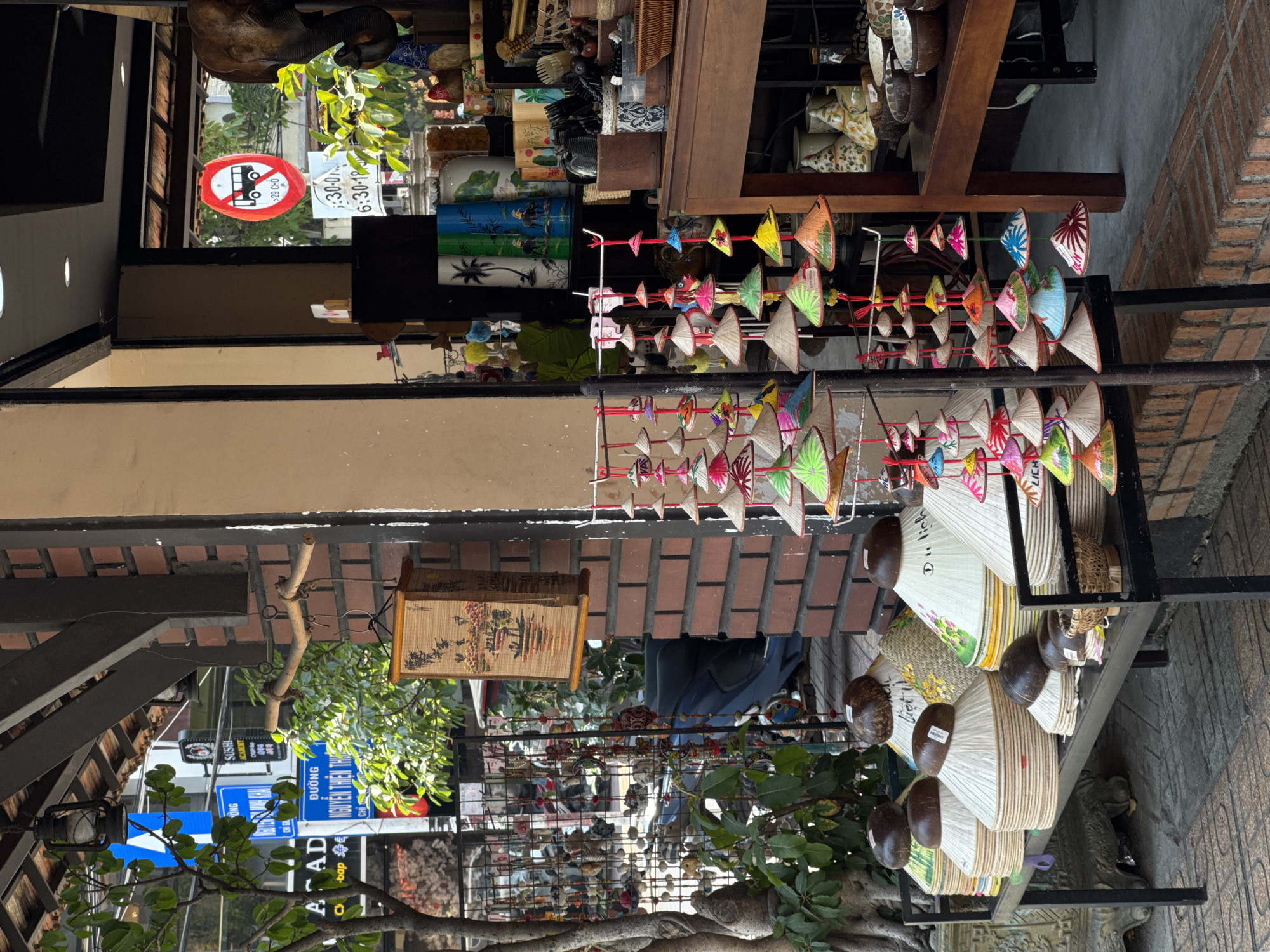 Captured outdoors in an urban setting, the photo presents an eye-level perspective of a street in Unknown, Unknown. The frame is dominated by a storefront, exhibiting a variety of souvenirs, including several miniature Vietnamese conical hats hung on strings. Additionally, there's an assortment of fans and other decorative items on display. In the background, street signs and various businesses are visible, indicating a bustling commercial area. The environment includes elements of nature such as trees and green foliage, which add vibrancy to the scene. People are discernable, though not the primary focus, with some appearing to walk past in the background. The architectural design of the buildings and the materials used (brick, concrete) suggest the local style and culture of the area. Overall, the image portrays a lively street scene with souvenir stalls typical of tourist destinations.
