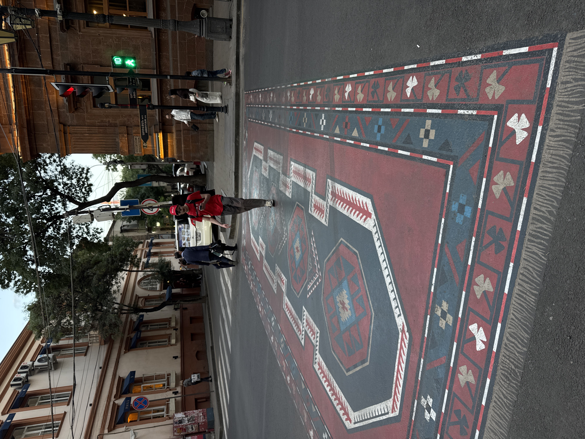 A street scene in Yerevan, Armenia shows a large, painted rug design on the asphalt of a crosswalk. The design is red, grey, and white with geometric patterns, resembling a traditional carpet. People are walking on and around the crosswalk. A man in a red shirt is standing on the rug design, gesturing with his hand. Cars are visible in the background. Buildings line the street with architectural styles that suggest a European influence. A traffic light is seen displaying a green walking signal for pedestrians. Street signs in Armenian script are also visible.