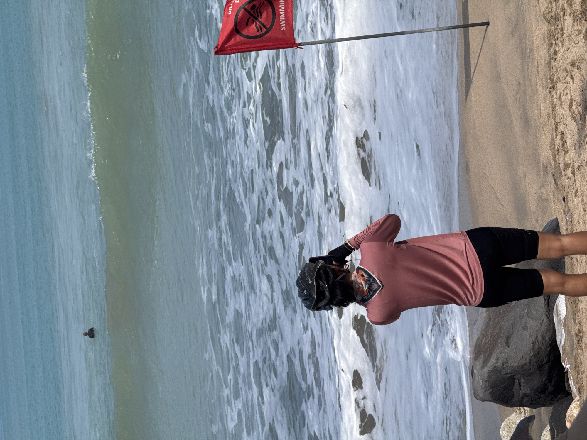 This image depicts a beach scene in Kuta, Indonesia. In the foreground, a person wearing a helmet, a pink long-sleeved shirt, and black shorts is standing on the sand, looking out at the ocean and holding a phone as if taking a picture or video. A red flag with a prohibition symbol indicating "no swimming" is planted in the sand to the right of the person. In the background, a person is visible in the water. The ocean is characterized by waves and foam. Large rocks are present on the beach in the foreground to the left.