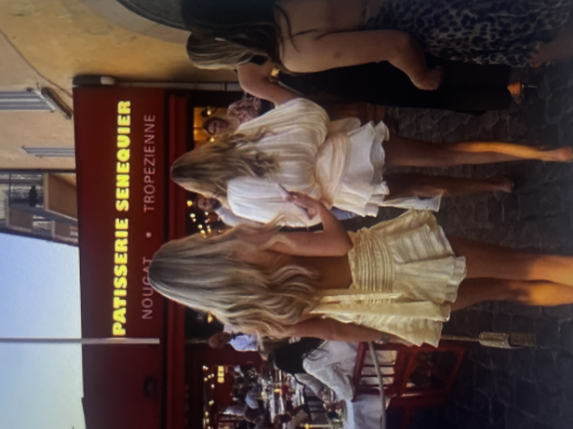 The image shows a group of women walking outdoors in front of a building with a sign that reads "PATISSERIE SENEQUIER NOUGAT TROPEZIENNE". The building appears to be a cafe or restaurant with outdoor seating. The women are dressed in summer attire. The environment suggests a warm climate, likely a tourist area. The sign indicates a location in Saint-Tropez, France, not Kołobrzeg, Poland.