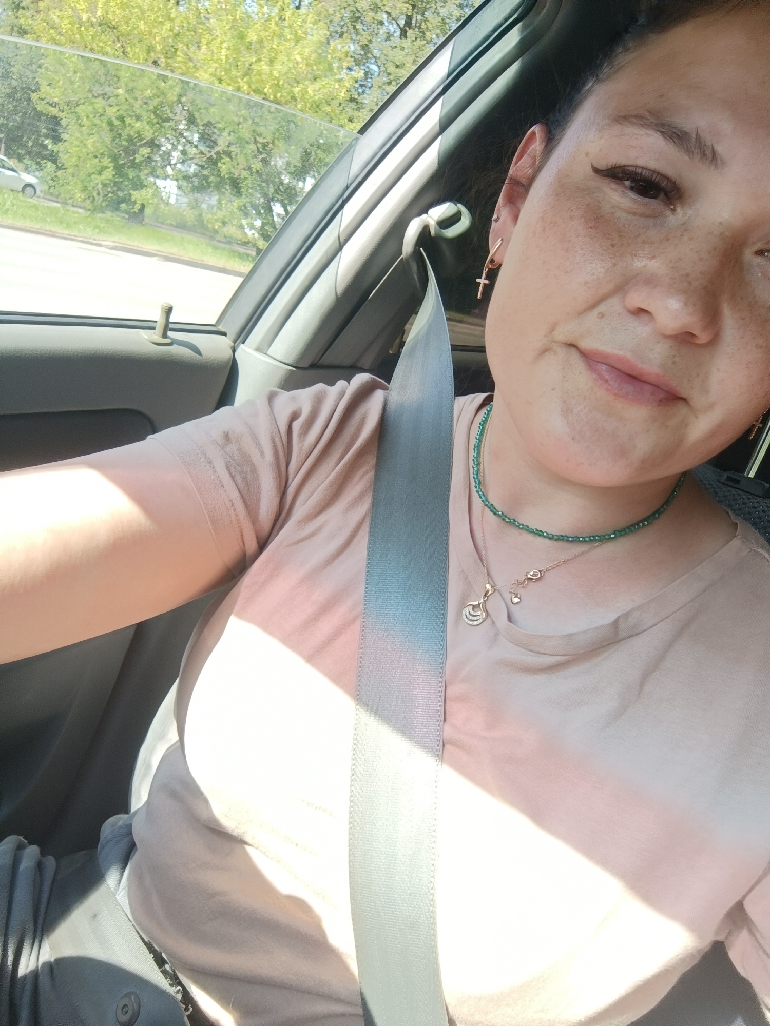 The image is a selfie taken inside a car. The person in the photo is a woman with dark hair, freckles, and is wearing a light-colored t-shirt, a green beaded necklace, and a silver necklace with a pendant. A seatbelt is visible across her chest. Sunlight streams into the car, creating shadows on her clothing. Outside the car window, trees and a street are visible, suggesting an outdoor setting. The image does not contain enough visual information to determine the specific location within the city of Chaikovsky, Russia.