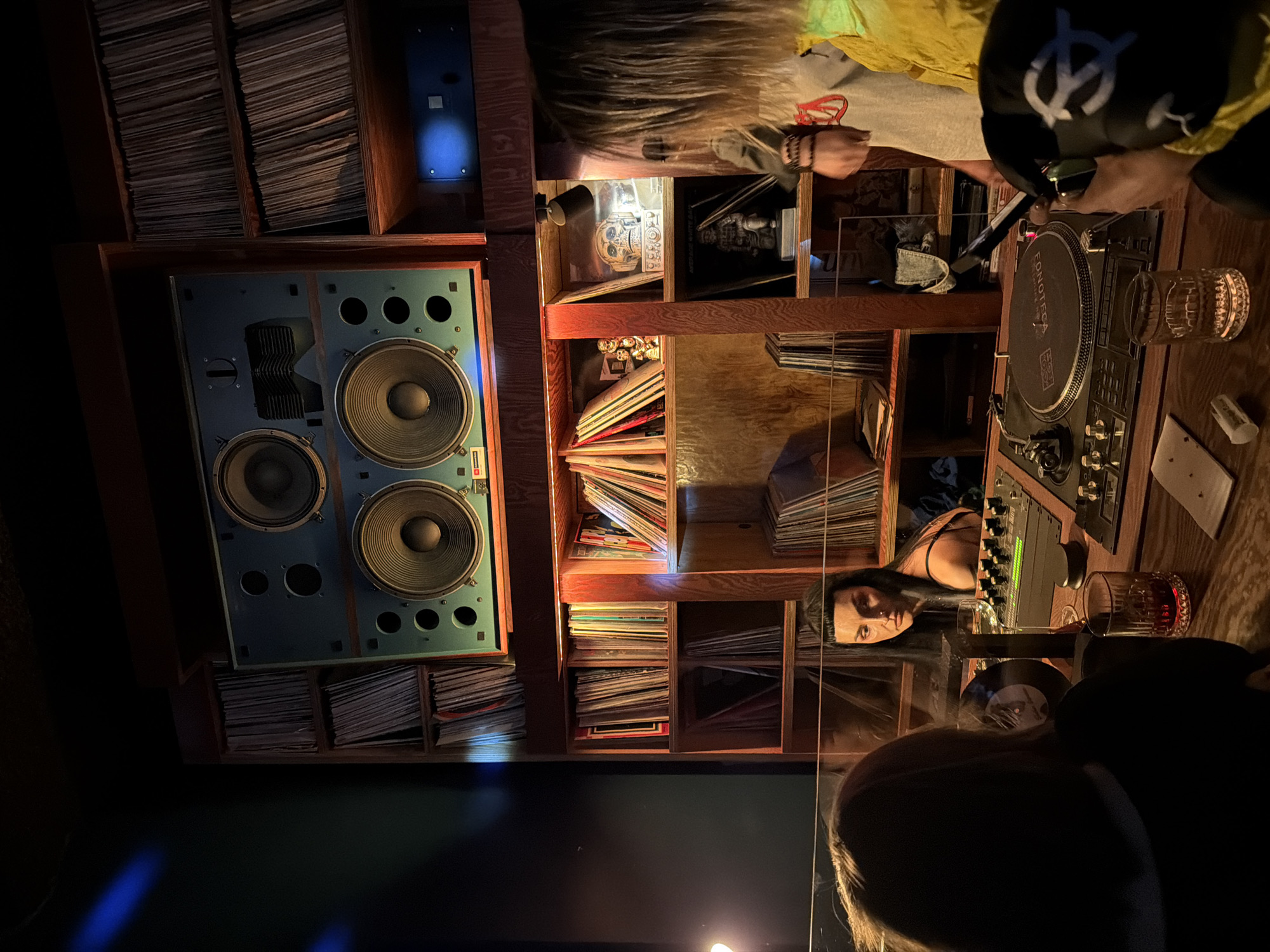 The image shows an interior scene with a focus on a DJ setup within a wooden shelving unit filled with vinyl records. A large speaker is positioned to the left of the shelving. At the top of the image, a person with long blond hair and another wearing a black baseball cap are interacting with the DJ equipment. Reflections indicate the presence of two women on the right side of the frame, looking towards the DJ setup. One is near the bottom, while the second is directly in front of the record player. A turntable and a mixer are prominent features, alongside a glass of liquid and other items. Based on the information available, a specific location in Moscow, Russia cannot be determined.