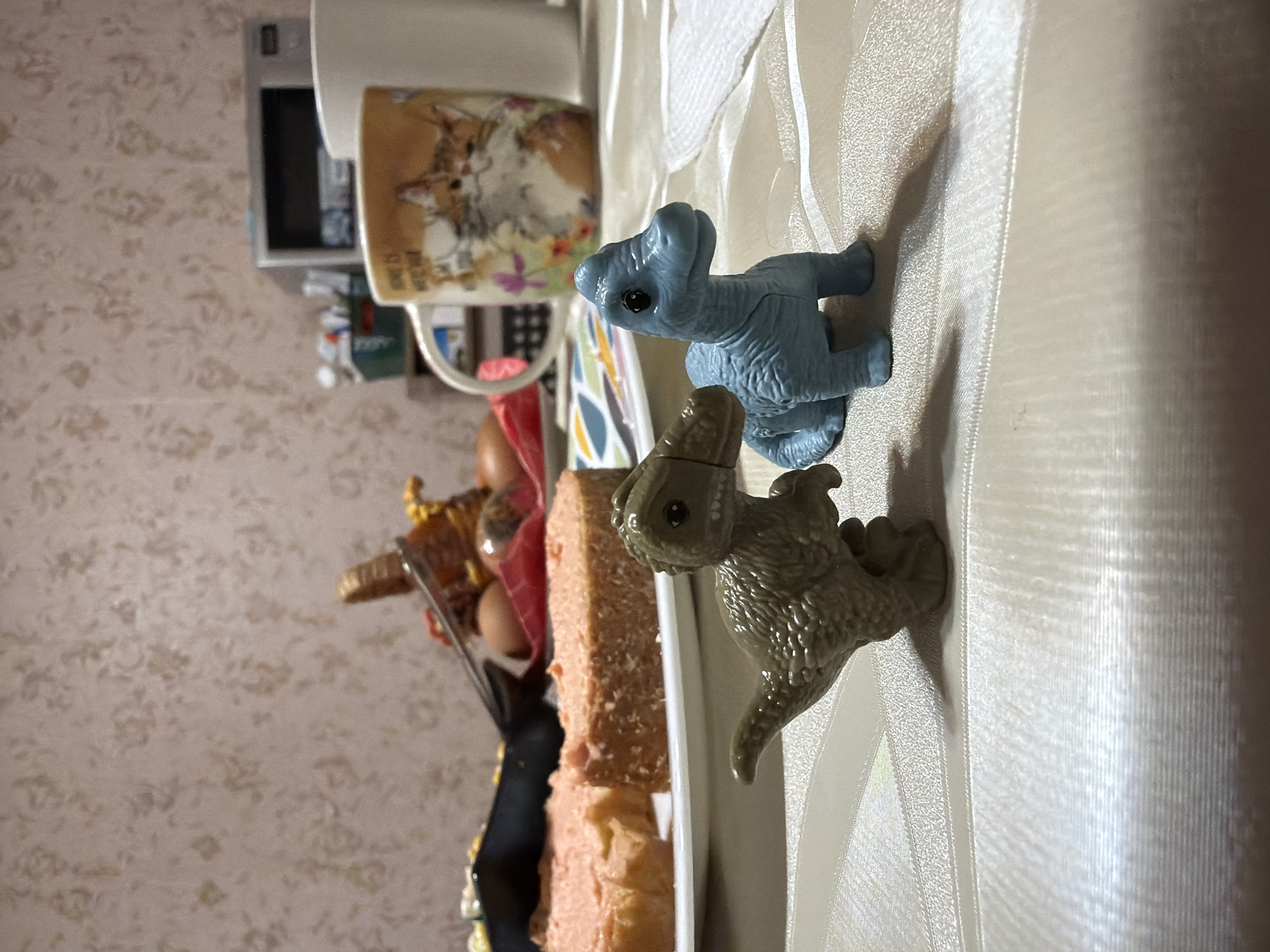 On a light-colored tablecloth, two toy dinosaurs, one blue and one olive, sit near a white rectangular dish. The dish holds a cake with multiple layers of light brown frosting. A cup with a cat illustration is next to the cake and dish. A tray filled with Easter eggs is slightly out of focus in the background. The location is Izhevsk, Russia.
