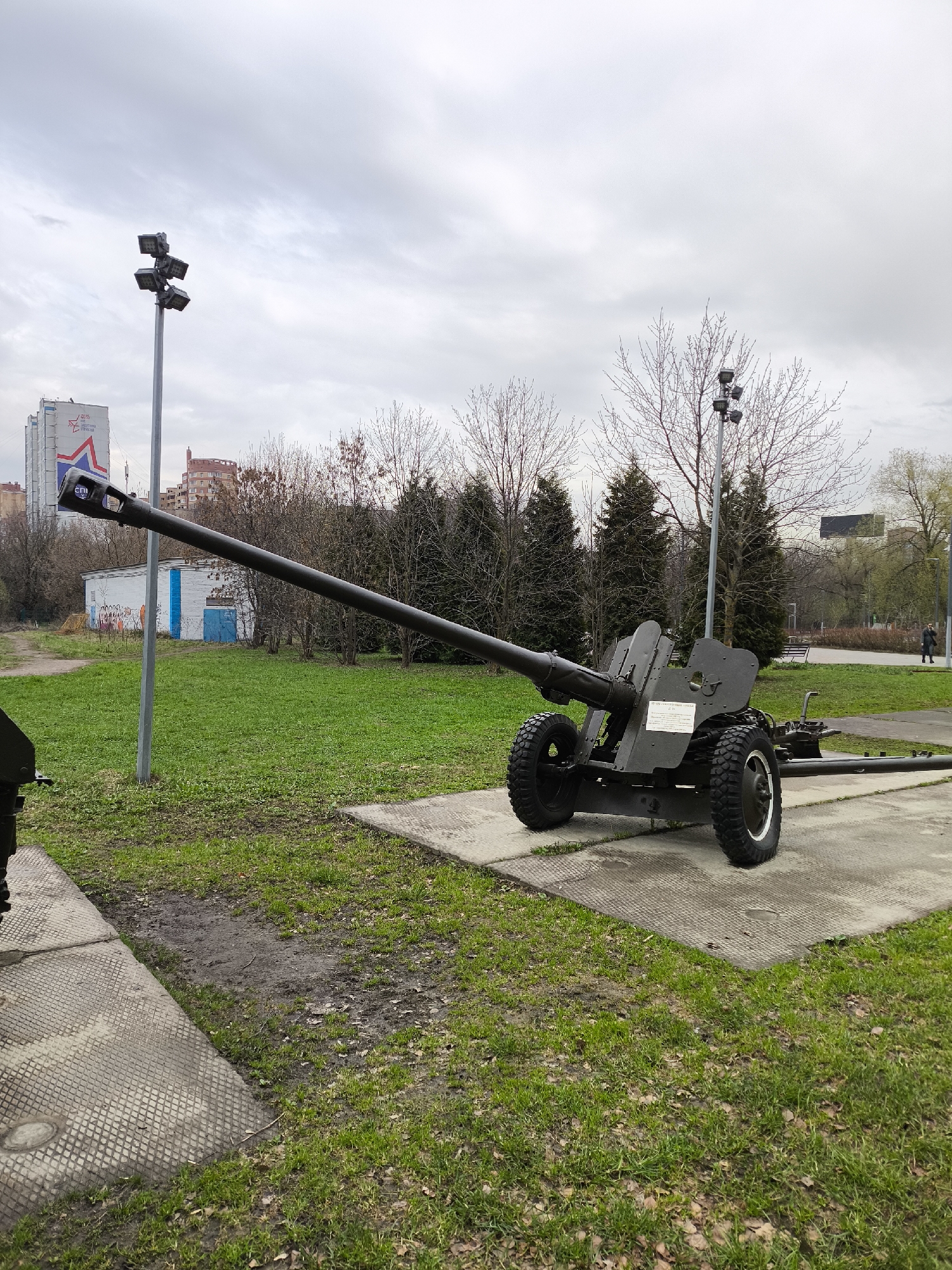The image captures an outdoor scene centered on a dark-colored 45-mm anti-tank gun, model 1937 (53-K), displayed as a monument or exhibit. The artillery piece, with its long barrel and large wheels, rests on a patterned concrete slab. A white plaque on the gun's shield provides specifications in Cyrillic: "45-мм противотанковая пушка образца 1937 г. (53-К), Калибр: 45 мм, Масса в боевом положении: 560 кг, Расчёт: 5 человек" (45-mm anti-tank gun, model 1937 (53-K), Caliber: 45 mm, Weight in combat position: 560 kg, Crew: 5 people).
The setting is an urban park or memorial area. The ground is covered with green grass interspersed with patches of dirt and concrete pathways. The sky is overcast, indicating a cloudy daytime, possibly in late winter or early spring, given the bare branches of many deciduous trees in the background. Some evergreen trees are also visible.
Two tall poles with multiple spotlights illuminate the area. In the background to the left, a large building displays a prominent red and white star logo and the Cyrillic text "ДТС" (DTS or GTS), with smaller text below possibly reading "ДОСААФ России" (DOSAAF Russia), suggesting a Russian location. Behind the gun's barrel, a smaller white building with blue vertical stripes and graffiti is visible. Further in the background, on the right, a solitary person walks along a paved path. No specific activity beyond viewing the monument is depicted.