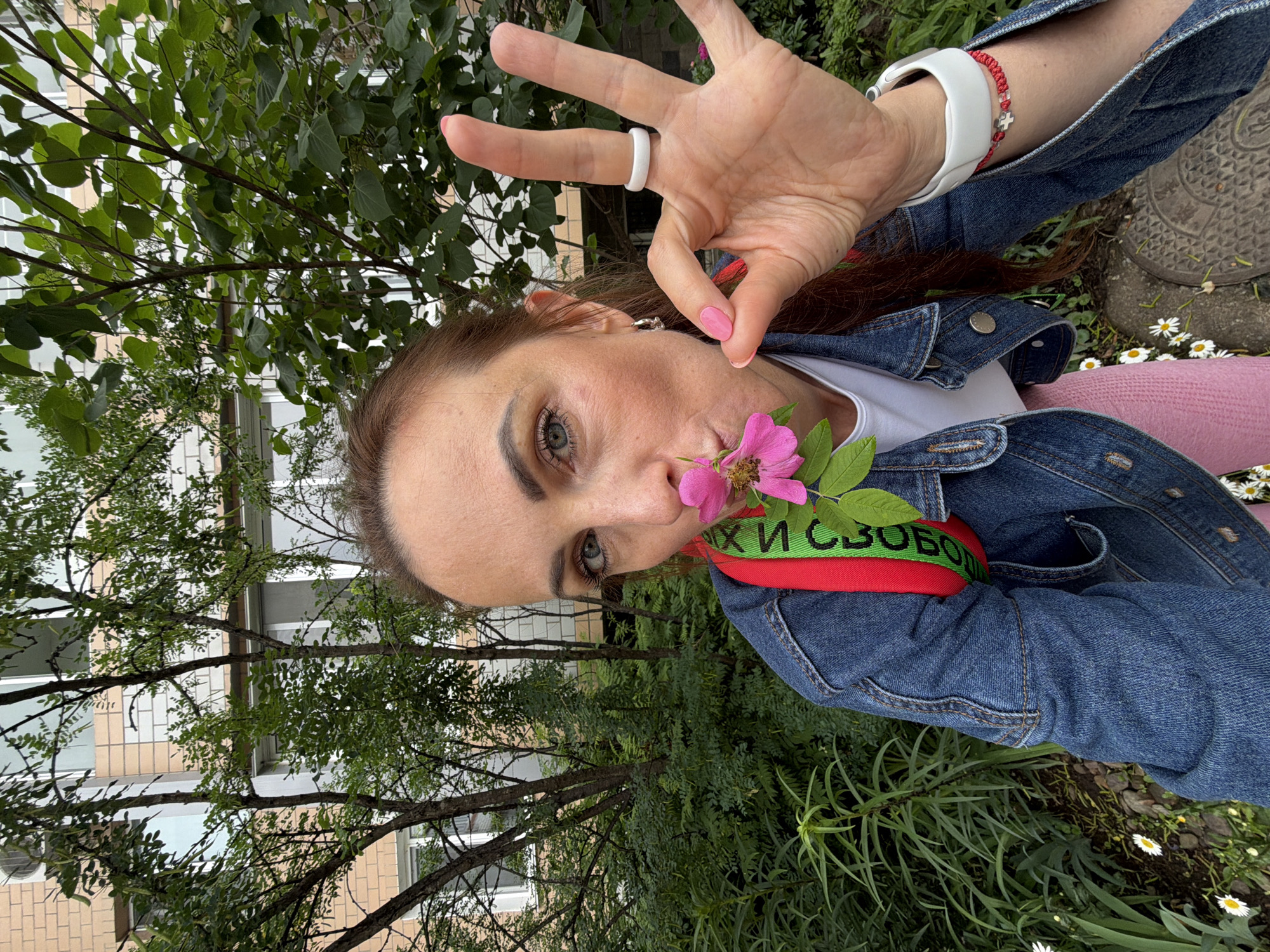 A medium shot shows a woman holding a pink flower to her nose and making an "okay" gesture with her right hand. She is wearing a denim jacket over a white t-shirt and a pink skirt. A red strap with green lettering is visible across her chest. She has light blue eyes and long brown hair pulled back. She is wearing a white ring on her right index finger and a white smartwatch with a red beaded bracelet on her left wrist. The background consists of lush green foliage, trees, and a building with light-colored brickwork and windows. Small white daisies are visible in the lower foreground, along with a manhole cover. The scene appears to be outdoors in a residential area, possibly in Birobidzhan, Russia, given the presence of the building and foliage typical of the region.