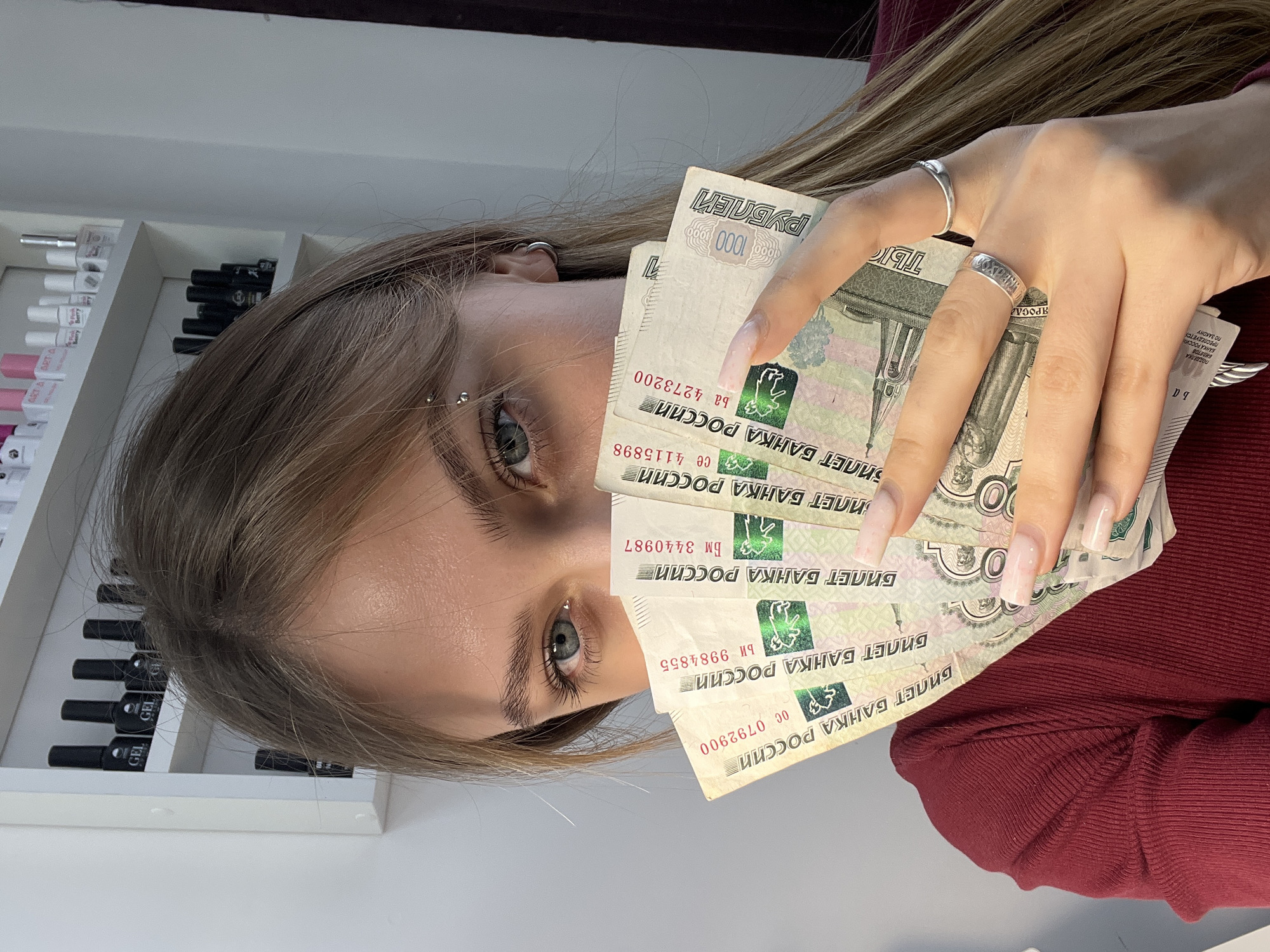 A young woman is holding a fan of Russian 1000 Ruble banknotes in front of her face. She has fair skin, light brown hair, and gray eyes with an eyebrow piercing. She wears two silver rings on her fingers. The banknotes are printed with the text "БИЛЕТ БАНКА РОССИИ" and "1000 РУБЛЕЙ". Behind her is a white shelf with rows of small bottles, some labeled "GEL". She is wearing a maroon-colored top. The background is blurry and indeterminate. The city of Фоки, Russia is not discernible in this image.