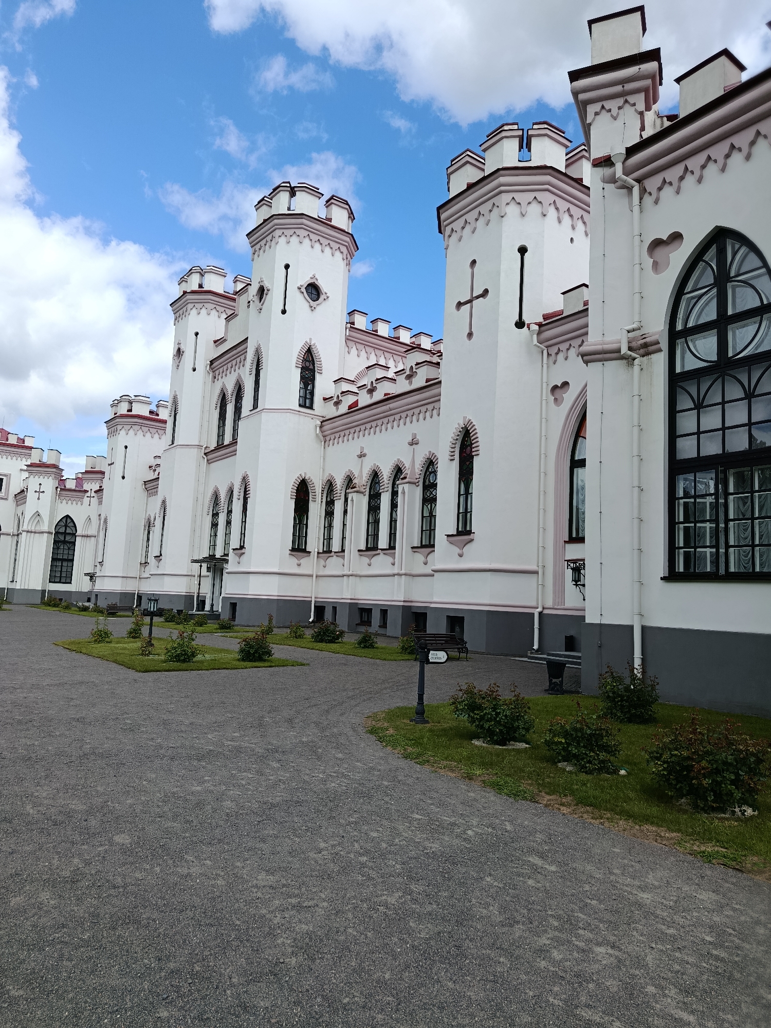 The image depicts a white, multi-turreted castle with pink accents under a partly cloudy blue sky. The castle features a series of pointed roofs, arched windows, and decorative crenellations. A paved pathway leads up to the building, with manicured green spaces and small bushes flanking it. Benches are visible near the building, and a lamppost stands on the pathway. The overall scene suggests a well-maintained historical building in an outdoor setting. The location is specified as Квасевіцкі сельскі Савет, Belarus. There are no people or animals visible in the image.
