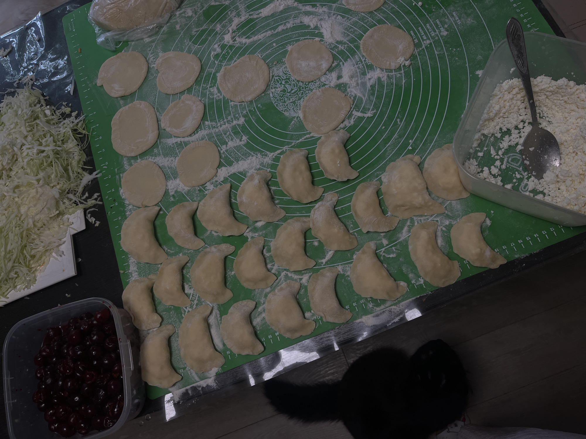 In Tikhoretsk, Russia, a dimly lit kitchen counter is the setting for food preparation. A large plastic container filled with dark red cherries sits to the left. To the right, a pile of shredded cabbage rests on a white surface. In the center, a green silicone baking mat is dusted with flour, displaying numerous rounds of uncooked dough and crescent-shaped pierogi. A black cat's silhouette is visible on the wooden floor to the left. Below the pierogi, a clear plastic container holds a mound of cottage cheese, with a spoon resting on top. The scene captures the process of making traditional Russian dumplings.