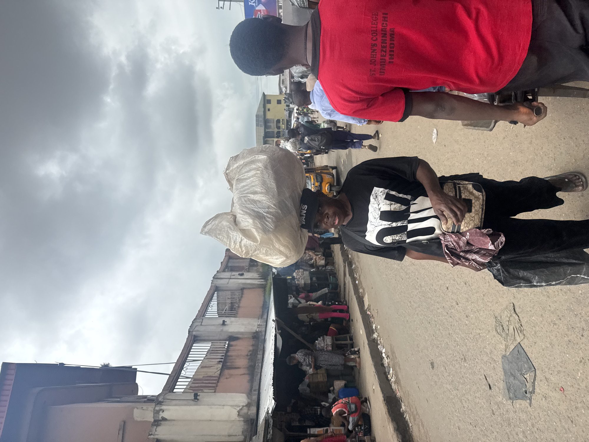 The image depicts a street scene in Nkpor, Nigeria, under an overcast sky. In the foreground, a person wearing a black t-shirt with "LVNS" visible across the chest carries a large, white, plastic-wrapped bundle on their head. They also have a small bag slung across their body and a black plastic bag in their hand. To the right, a person in a red t-shirt with "ST. JOHN'S COLLEGE UMUEZENNACHI ENUGU" printed on the back is also visible. The street is paved and appears to be a market area, with stalls and shops lining the left side of the road. Several other individuals are present in the background, along with a yellow tricycle. The environment suggests a bustling commercial area with a somewhat unkempt appearance, as evidenced by debris on the street.