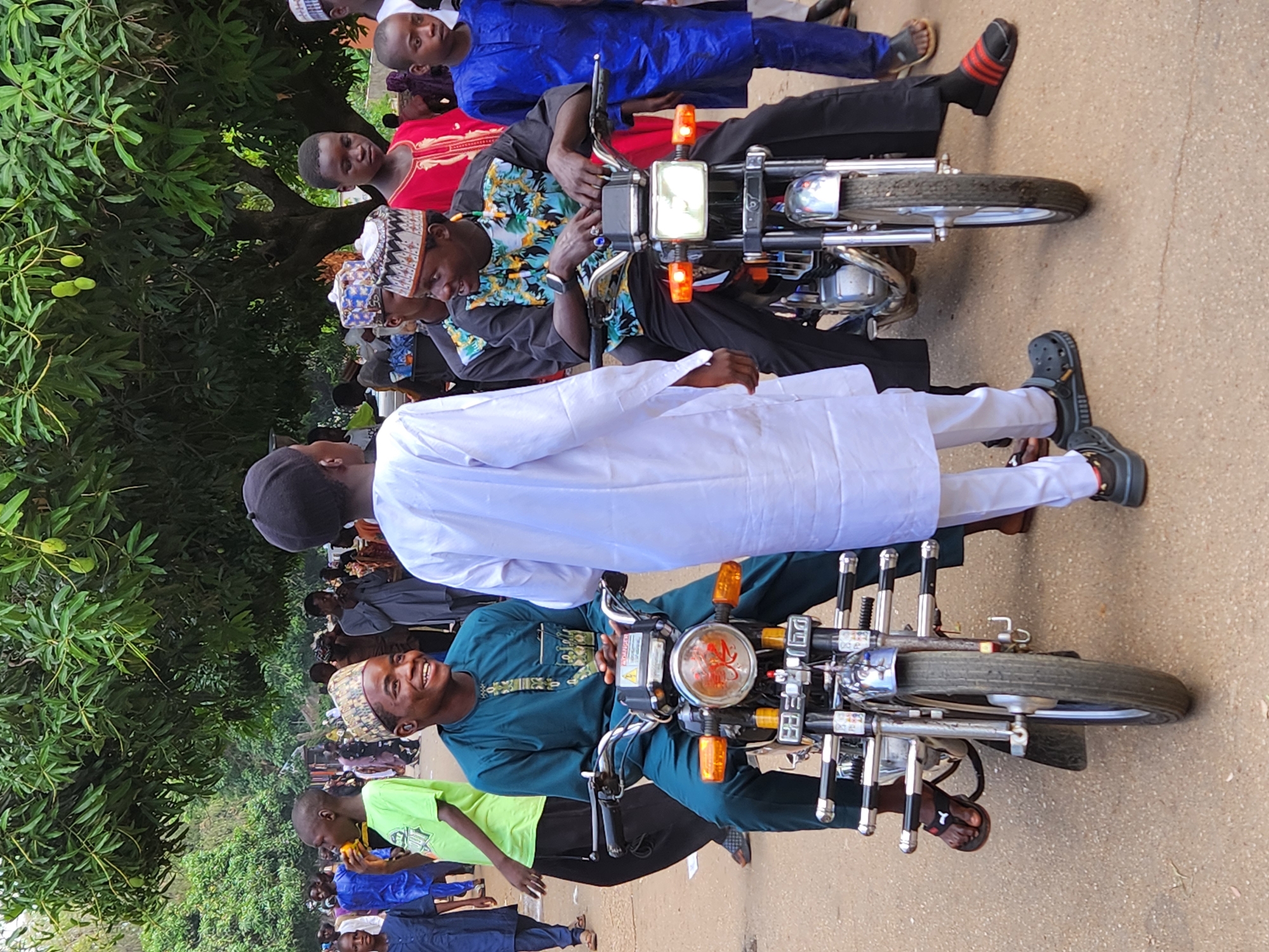 The image shows a group of people in what appears to be a street setting. Several individuals are sitting on motorcycles, while others are standing nearby. The individuals are dressed in various styles of clothing, with some wearing traditional garments. The motorcycles have classic designs. The background includes trees and possibly other buildings. The location is determined as Jos, Nigeria.