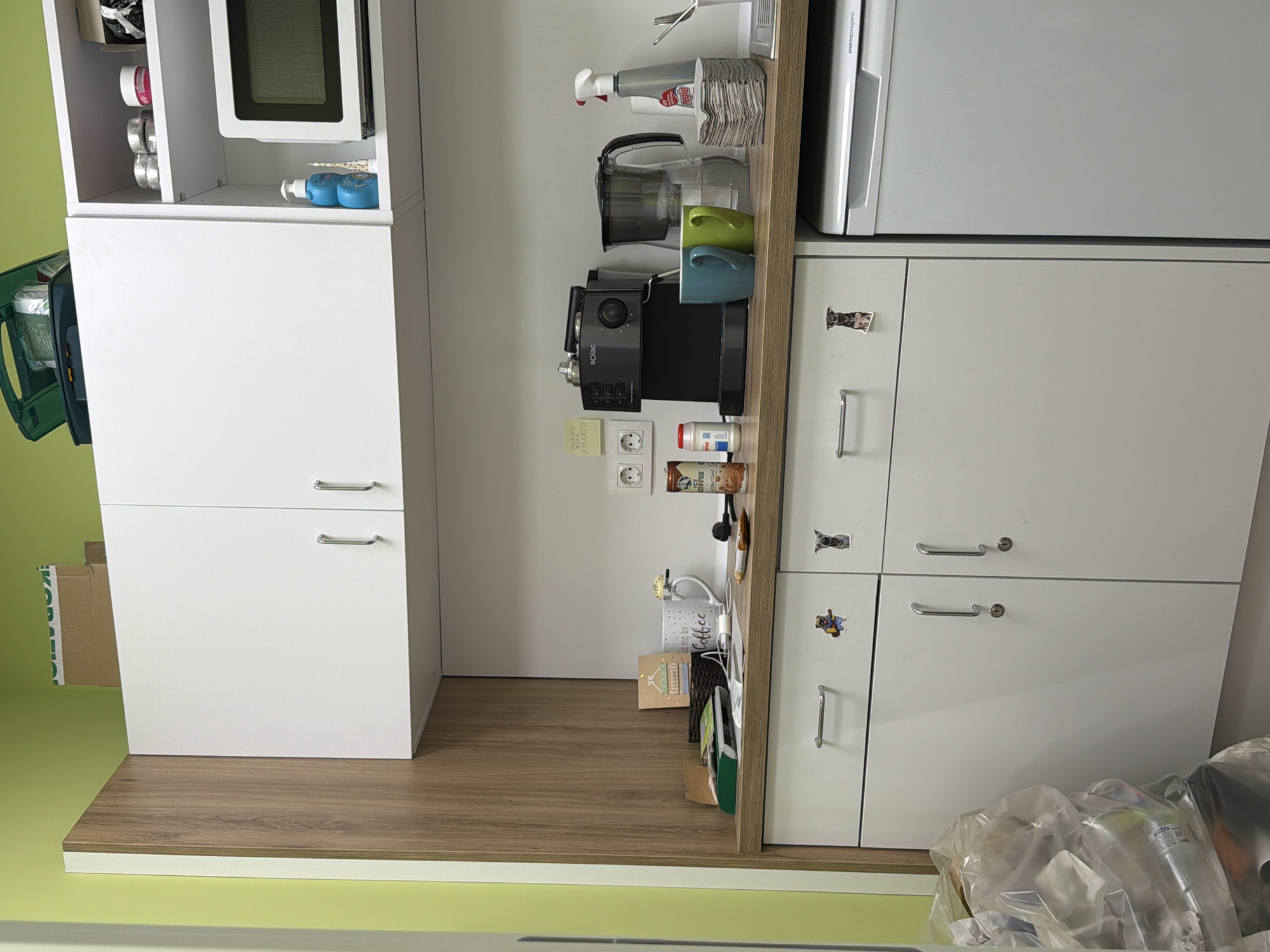 The image shows a kitchen interior with white cabinets and drawers on a brown wooden floor, located in Sofia, Bulgaria. On the left, a tall white cabinet with a microwave and shelving above, and a two-door cabinet below. The cabinet is adjacent to a vertical storage unit with a coffee machine, cleaning supplies, and mugs. To the right is a refrigerator placed on top of two rows of cabinets with silver handles and animal-themed stickers. Various items are stored in the cabinet, while transparent plastic bags are visible on the floor in front of the right cabinet. The walls are painted in a light green color.