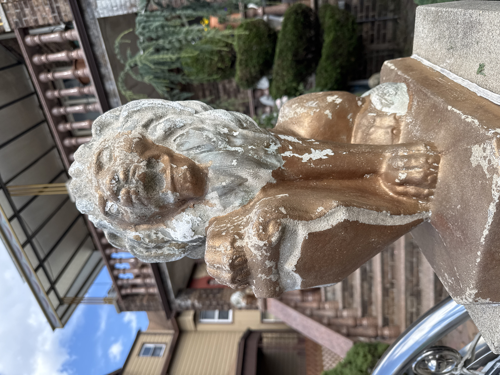 A weathered, gold-painted lion statue sits prominently in the foreground of this outdoor, daylight scene, likely in a residential area of New York City, United States of America. The statue, which appears to be made of concrete or a similar material, exhibits significant wear and tear, with large areas of paint chipped away to reveal a lighter-colored material underneath. Its features, including its mane, face, and paws, are visible but softened by age and exposure. The lion is posed in a seated position, with one paw resting on what appears to be a smaller, cub-like figure, also weathered.
Behind the statue, a multi-story house with a beige exterior and brown trim is visible. A covered porch with a metal roof and decorative columns is present, as is a set of stone stairs leading up to the house. To the right of the house, an elaborately shaped green bush, possibly a topiary, stands tall. Further in the background, a tall, leafy tree with cascading branches adds to the natural elements of the scene. The sky above is a mix of blue and white clouds. A metallic railing, possibly part of a fence or staircase, is visible in the lower left corner of the image. The overall impression is one of an older, possibly once grand, residential property with decorative architectural elements and landscaping.
