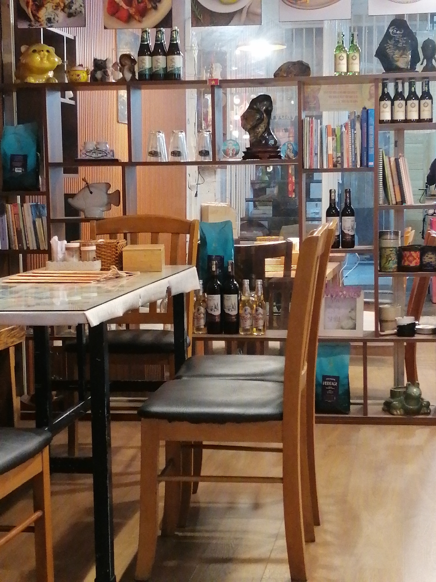 The image shows a cafe interior in Da Lat, Vietnam. The cafe features wooden shelves filled with books, bottles, and decorative items. A table with a patterned tablecloth is set with placemats and cutlery. Wooden chairs surround the table, and some are empty. In the background, more shelves are visible, stocked with various objects. The lighting is soft, creating a cozy ambiance. The scene appears to be a quiet moment in the cafe, with no customers or staff actively engaged in the frame.