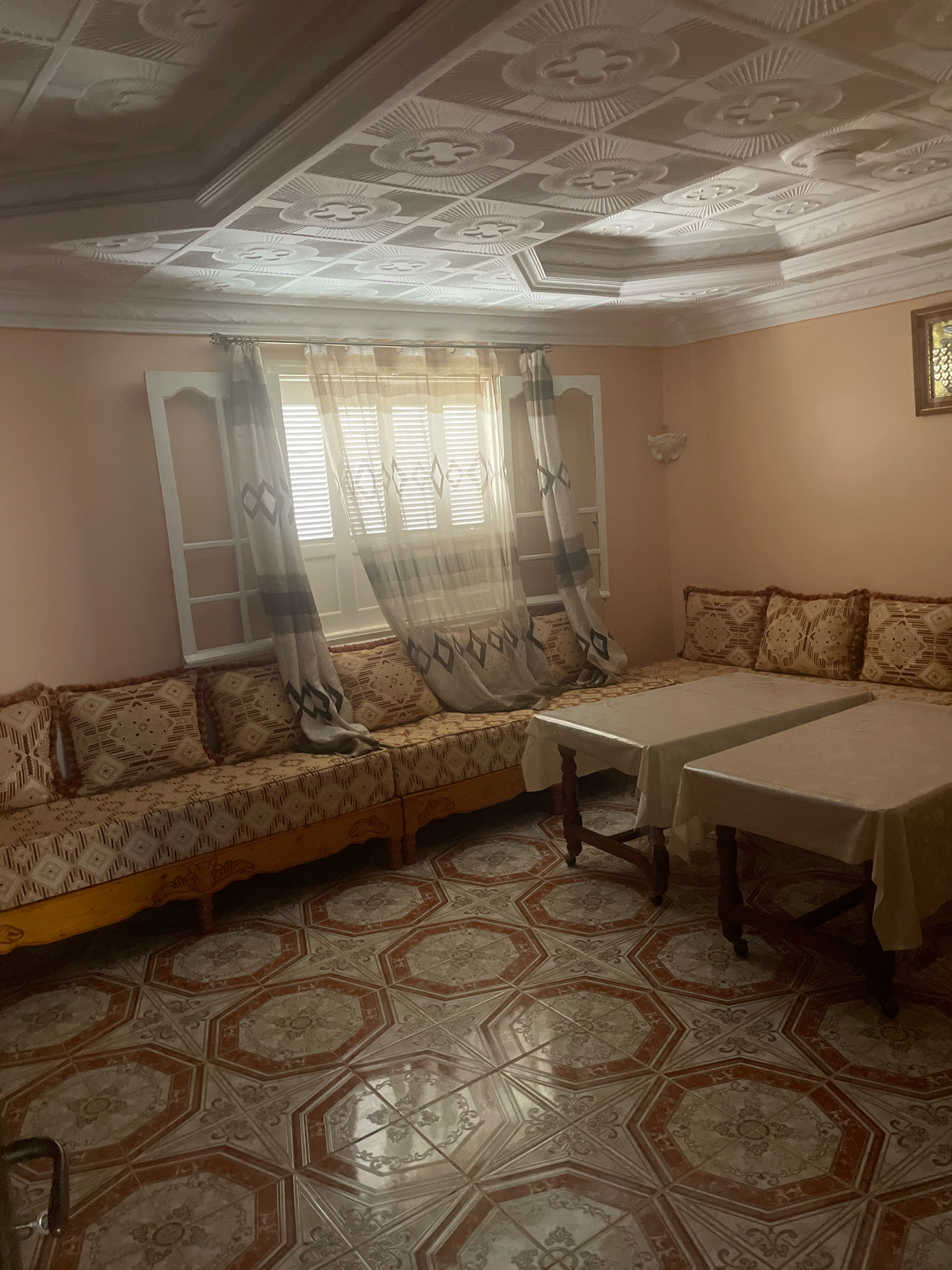 The image depicts an interior view of a room, likely a living room or reception area, in Ghazaouet, Algeria. The room features traditional Moroccan or Algerian design elements. The walls are painted a soft peach color, and the ceiling is adorned with intricate white plasterwork featuring geometric and floral patterns, divided into square panels.
The floor is covered with patterned tiles, exhibiting an octagonal design with a red border and a central motif that appears to be a stylized floral or crest.
A long, L-shaped sofa upholstered in a patterned fabric with geometric designs in shades of beige and brown lines the walls. It is adorned with matching throw pillows. In front of the sofa, two low wooden tables are placed side-by-side, each covered with a plain white tablecloth.
To the left, a large window is dressed with sheer curtains featuring a diamond pattern in gray and beige. The window itself has white mullions forming a grid. On the wall above the sofa, to the right, a small, ornate gold-framed picture or mirror is visible. A white wall sconce is mounted on the wall between the window and the picture.
The lighting appears to be ambient, likely from an artificial source not directly visible, casting soft shadows. The overall impression is that of a comfortable, traditional seating area. There are no people visible in the image.