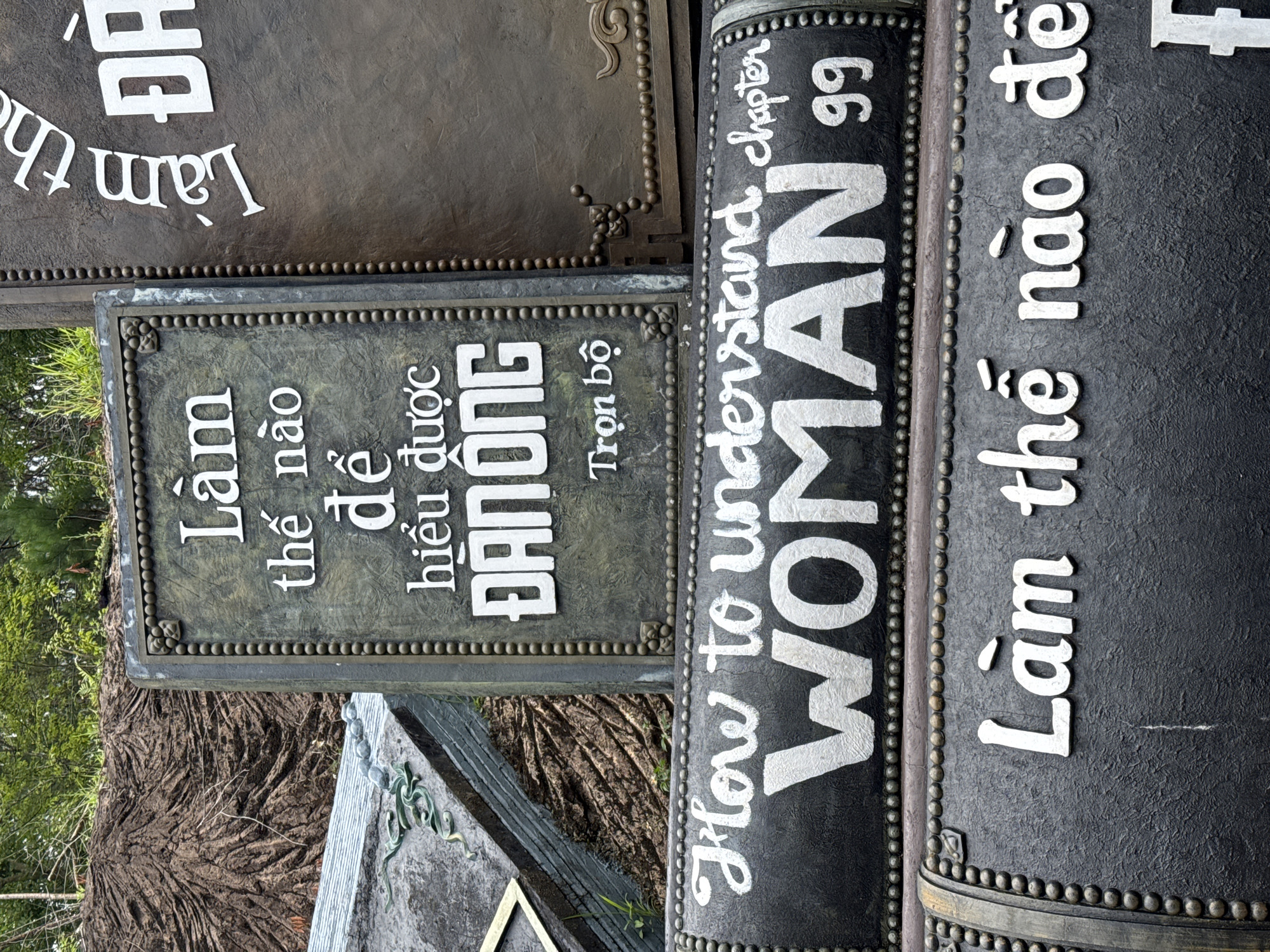 The image shows a collection of textured, dark-colored objects with white lettering, resembling a display or monument. These objects appear to be stacked or arranged vertically. The text is in both Vietnamese and English. One object has the Vietnamese phrase "Làm thế nào để hiểu được ĐÀN ÔNG Trọn bộ," which translates to "How to understand MEN Complete set." Another object features the English phrase "How to understand chapter WOMAN 99." The background shows greenery and what looks like a large, textured tree trunk or root structure, suggesting an outdoor setting, possibly in a park or garden. The lighting appears natural, suggesting it was taken during the daytime. The overall presentation is striking due to the contrast between the dark surfaces and the bright white text. The style of the lettering and the textured materials suggest an artistic or decorative purpose.
