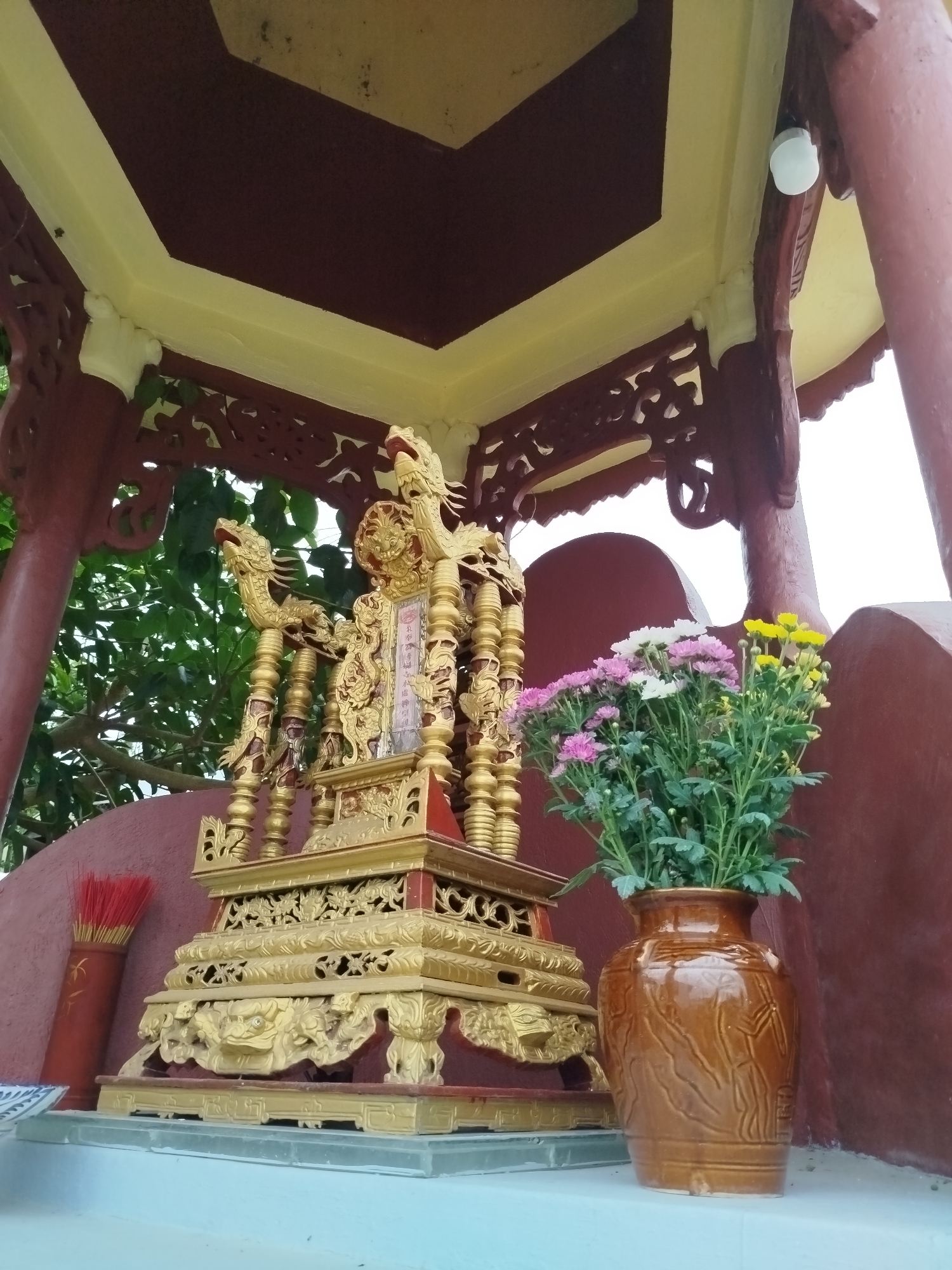 The image depicts an outdoor altar in Da Lat, Vietnam, characterized by a traditional Vietnamese architectural style. The altar is adorned with golden, intricately carved ornaments, including dragon motifs. In front of the altar, a vase of colorful chrysanthemums, featuring shades of pink, white, and yellow, adds a vibrant touch to the scene. To the left of the altar, a bundle of red incense sticks is arranged in a decorative holder, indicating a place of worship or remembrance. The background features lush green foliage, suggesting the altar is situated in a garden or natural setting. The overall ambiance is one of tranquility and reverence.