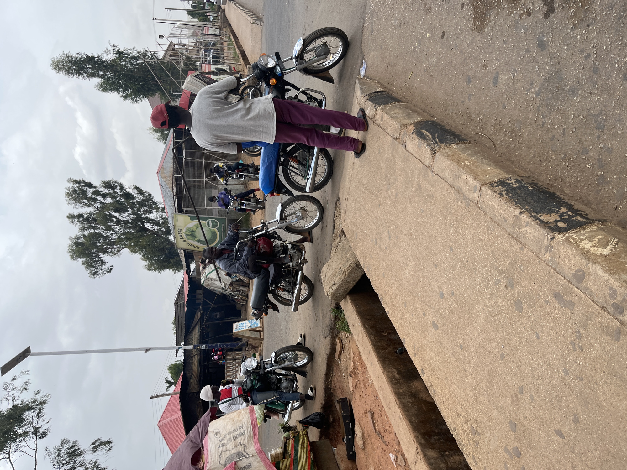 The image depicts a street scene in Bar Jirgi Summa, Nigeria. Several motorcycles are parked along the roadside, with individuals standing or sitting on them. A man wearing a red cap, grey shirt, and purple pants stands next to one of the motorcycles, seemingly engaged in a conversation. Behind the motorcycles, there are buildings and structures, including what appears to be a market area with stalls and signage. The sky is overcast, suggesting a cloudy day. The road is paved and has a concrete curb.