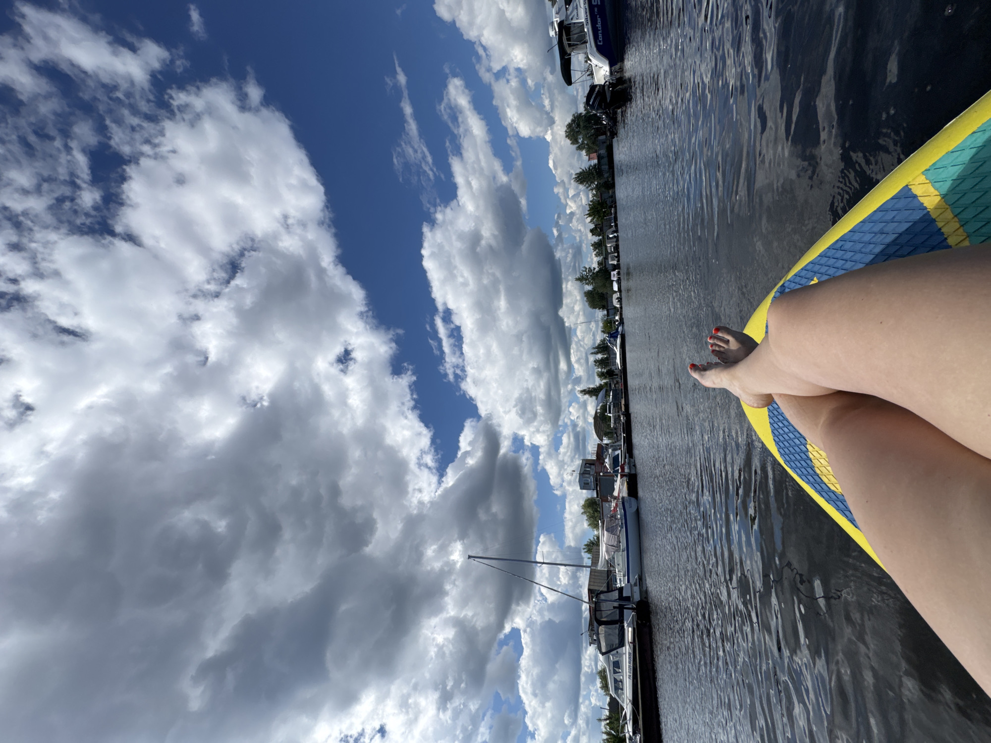An overhead perspective shot shows a person's legs from the knees down, resting on a yellow and blue paddleboard. The person's feet, with red painted toenails, are visible at the top of the frame. The paddleboard is on a body of water, which is dark and reflective, with ripples on the surface. In the background, a marina is visible with several boats docked along the shore. The boats vary in size and type, including sailboats and motorboats. Some of the boats have covers or Bimini tops. Behind the boats, trees and buildings are visible. The sky above is a bright blue with scattered white and gray clouds, suggesting a partly cloudy day. The scene appears to be set in a marina in the city of Chaikovsky, Russia, given the context of the marina and the boats.