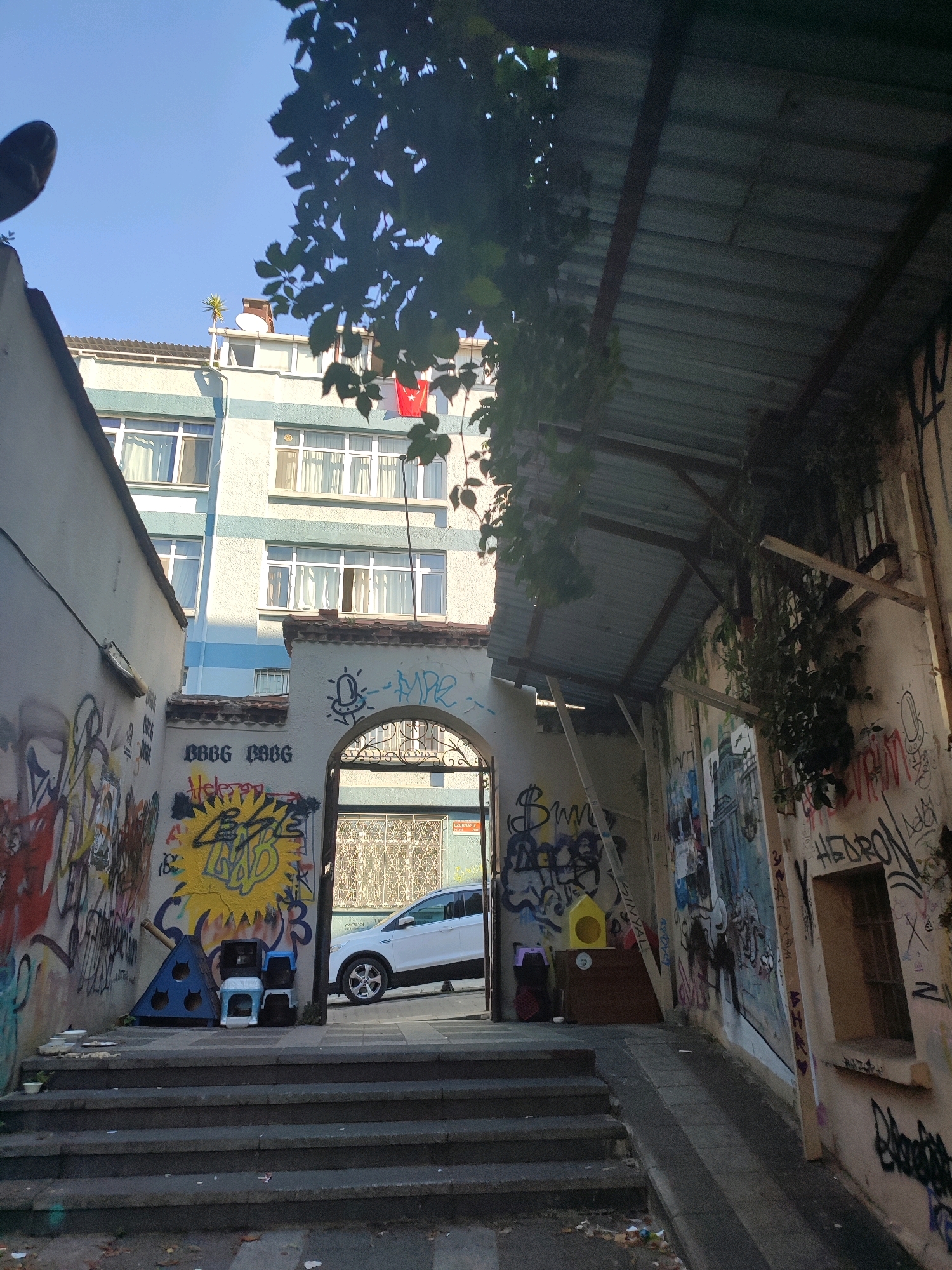 The image depicts an urban scene in Kadıköy, Turkey. In the foreground, a set of concrete stairs leads up to an arched entryway. The walls on either side of the entryway are covered in graffiti. To the left, a light blue building with white trim and multiple windows rises in the background. A white SUV is parked on an incline to the right of the entryway, partially visible. Overhanging the scene from the right is a corrugated metal roof supported by wooden beams, with vines growing down its sides. The overall impression is one of a somewhat dilapidated but vibrant urban environment, characteristic of many city districts.