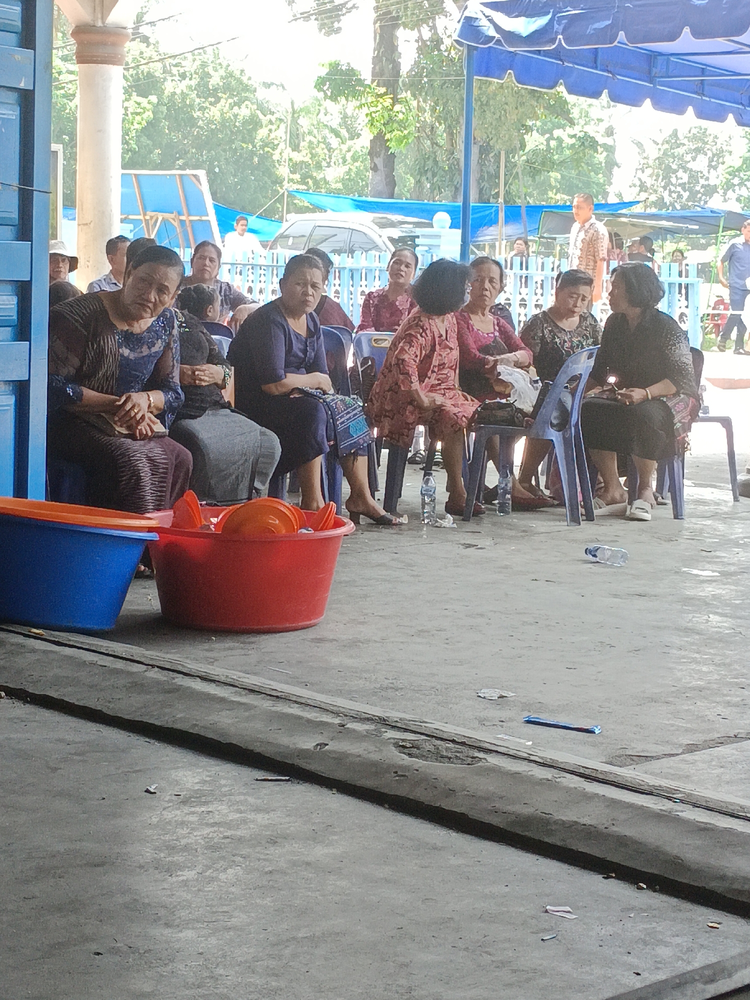 A group of people are seated on chairs outdoors, some of them wearing traditional clothing. Two large plastic tubs, one blue and one red, are in the foreground. The red tub is filled with orange dishes. The ground is concrete, and there is some debris scattered around. In the background, there are trees and a blue awning. The scene appears to be in a public area, possibly a gathering or event. The location is identified as Aek Kanopan, Indonesia.