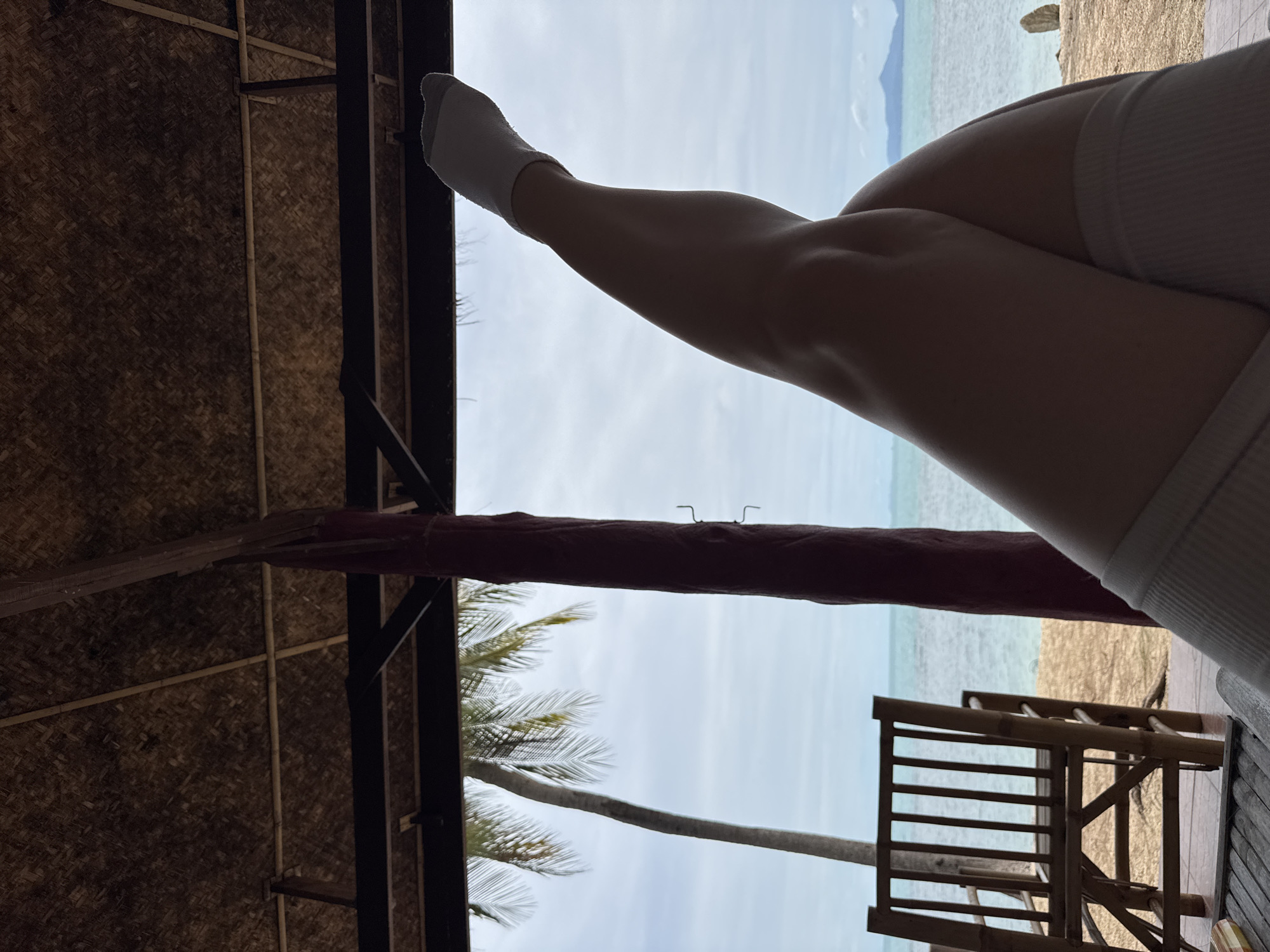 In Baan Tai, Thailand, a person's legs, clad in white socks, are positioned with one leg crossed over the other, in the foreground. The legs are silhouetted against a backdrop of the ocean, a sandy beach, and a palm tree. A bamboo chair is visible to the left, and part of a thatched roof structure is above. The scene appears to be from a covered outdoor seating area overlooking a beach.