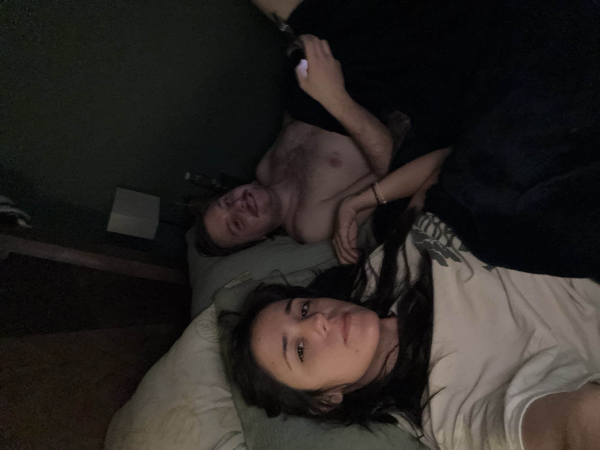 Two people are lying on a bed. A woman with long dark hair is lying on her side facing the camera, covered with a white sheet. A man is lying on his back above her, shirtless and holding a smartphone. The scene is dimly lit. The location is undetermined, and the city of Huixquilucan de Degollado, Mexico cannot be verified from the image.