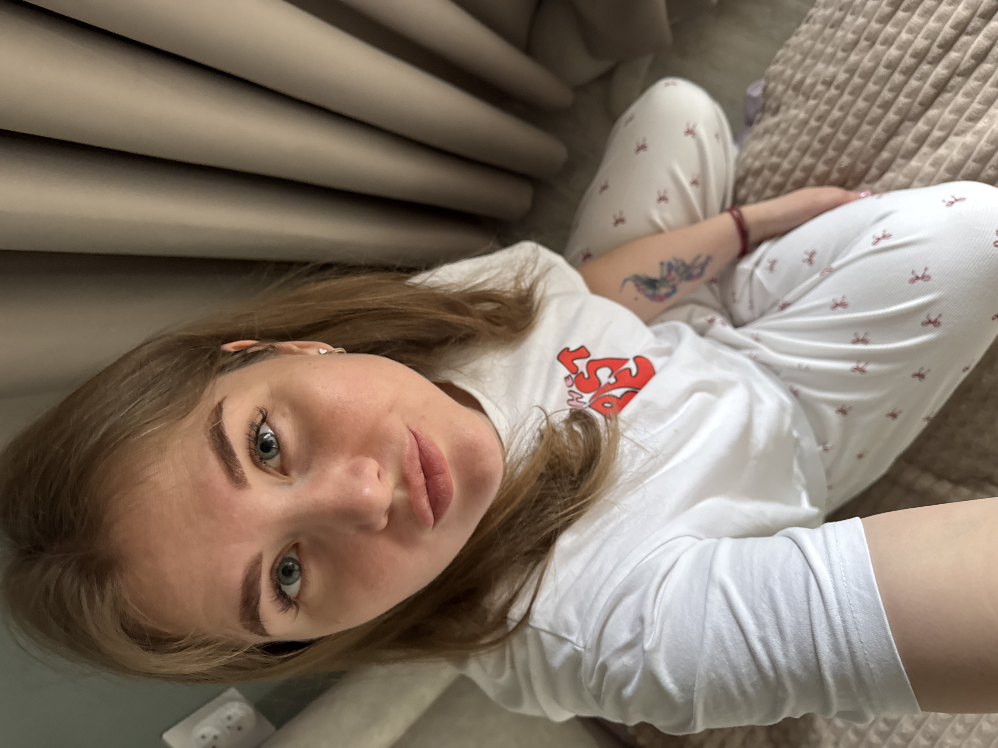 A young woman with light brown hair and blue eyes is looking at the camera. She is wearing a white t-shirt with a red logo and white pajama pants with a red bow pattern. She has a tattoo on her left arm. She appears to be sitting on a bed or couch with a textured beige cover. In the background, there is a light beige curtain. The location is likely Gornyi (Dvurechenskoe S/P), Russia.