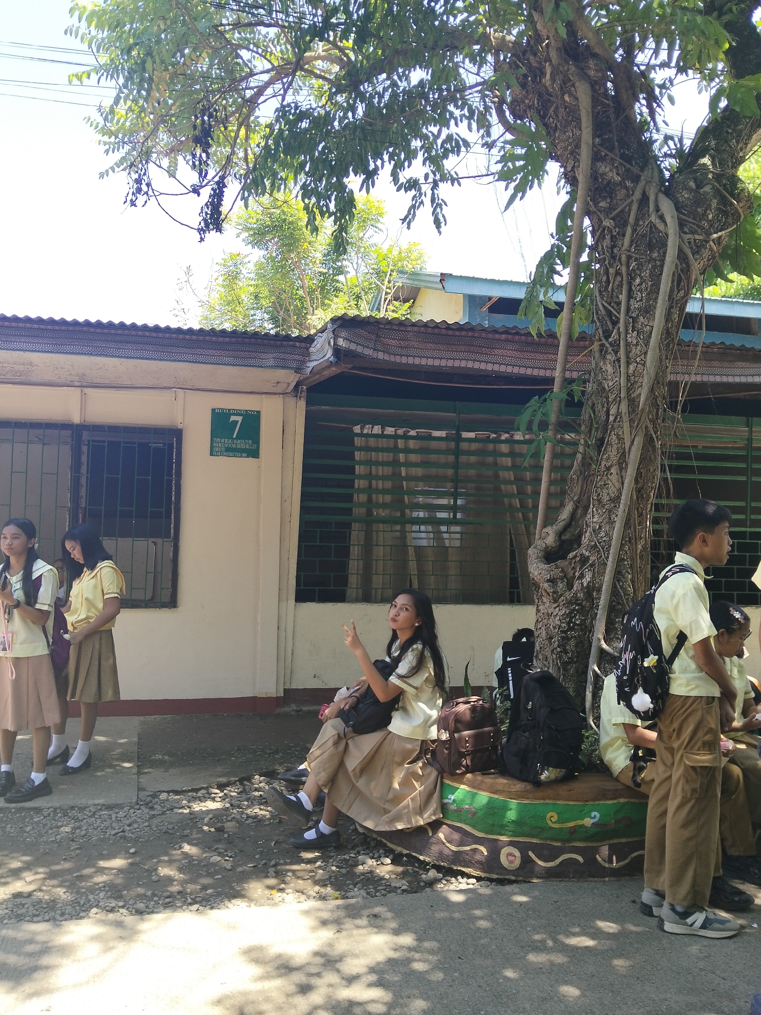 This is a medium shot photograph taken outdoors on a sunny day, likely in the Philippines, given the context of Davao City. The scene depicts a group of young students, appearing to be in uniform, gathered outside a building. The building itself is a light-colored, single-story structure with a corrugated metal roof and barred windows. A sign on the building reads "BUILDING NO. 7 TYPE OF BLDG MARCOS TYPE SOURCE OF FUND: DEPED BUGET YEAR CONSTRUCTED: 1994".
Several students are visible, predominantly female, wearing light yellow tops and beige or tan skirts, with white socks and dark shoes. One student is sitting on a decorative, colorful bench or planter, looking towards the camera with a slight smile and holding up two fingers in a peace sign. Other students are standing nearby, some appearing to be chatting or waiting. Backpacks are placed on the ground and on the bench. A large, mature tree with thick, gnarled branches and visible aerial roots stands prominently to the right of the building, casting dappled shadows on the ground. The bright sunlight and clear sky suggest it is midday. The overall atmosphere appears relaxed and casual, with the students seemingly taking a break or waiting for something.