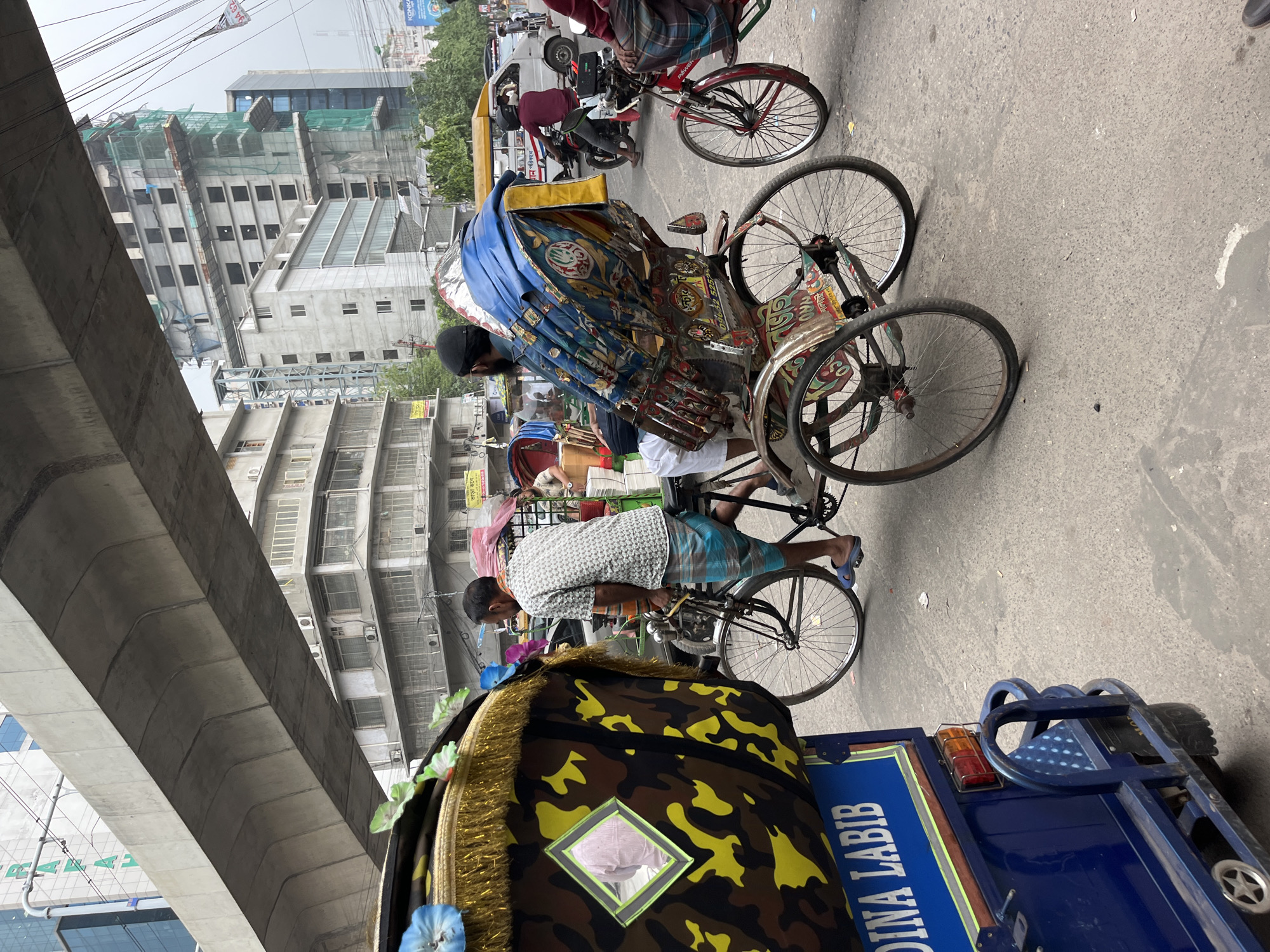The image depicts a street scene in Dhaka, Bangladesh. Two rickshaws, one with a blue canopy and the other with a yellow camouflage design, dominate the foreground. Rickshaw pullers are visible, one with a black cap and the other with a gray shirt, operating their vehicles. Other motorized vehicles are present, including a red motorcycle and a blue auto-rickshaw. The urban backdrop includes multi-story buildings, some under construction, and an elevated concrete structure. Overhead electrical wires crisscross the sky.