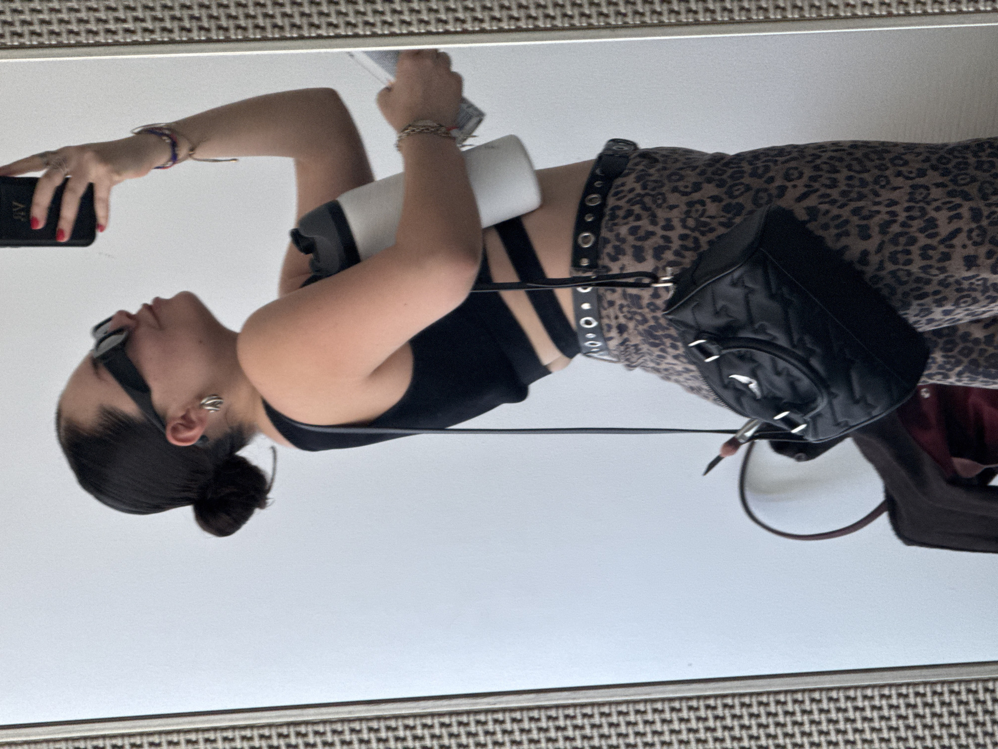 A young person, likely female, is depicted taking a mirror selfie in an indoor setting, presumably in Mexico City, Mexico. The subject is seen from the chest up, facing to her left, with her head slightly tilted upwards.
She has dark hair pulled back into a bun and is wearing black sunglasses. A silver-toned ear cuff adorns her visible right ear. Her attire includes a black crop top featuring multiple straps, paired with distinctive leopard print pants. A black belt with silver grommets and a silver buckle cinches her waist.
In her right hand, which displays red-painted fingernails, she holds a black smartphone that captures the reflection. Partial text "RY" is visible on the phone case. Her left arm cradles a white and black insulated bottle or tumbler. A black quilted shoulder bag with silver hardware hangs from her right side, while a second, larger, dark-colored bag is partially visible hanging lower on the right.
The background reflects a plain, light-colored wall or surface. A patterned, textured border, possibly the mirror frame, runs along the top of the image. The overall impression is a posed, casual snapshot.