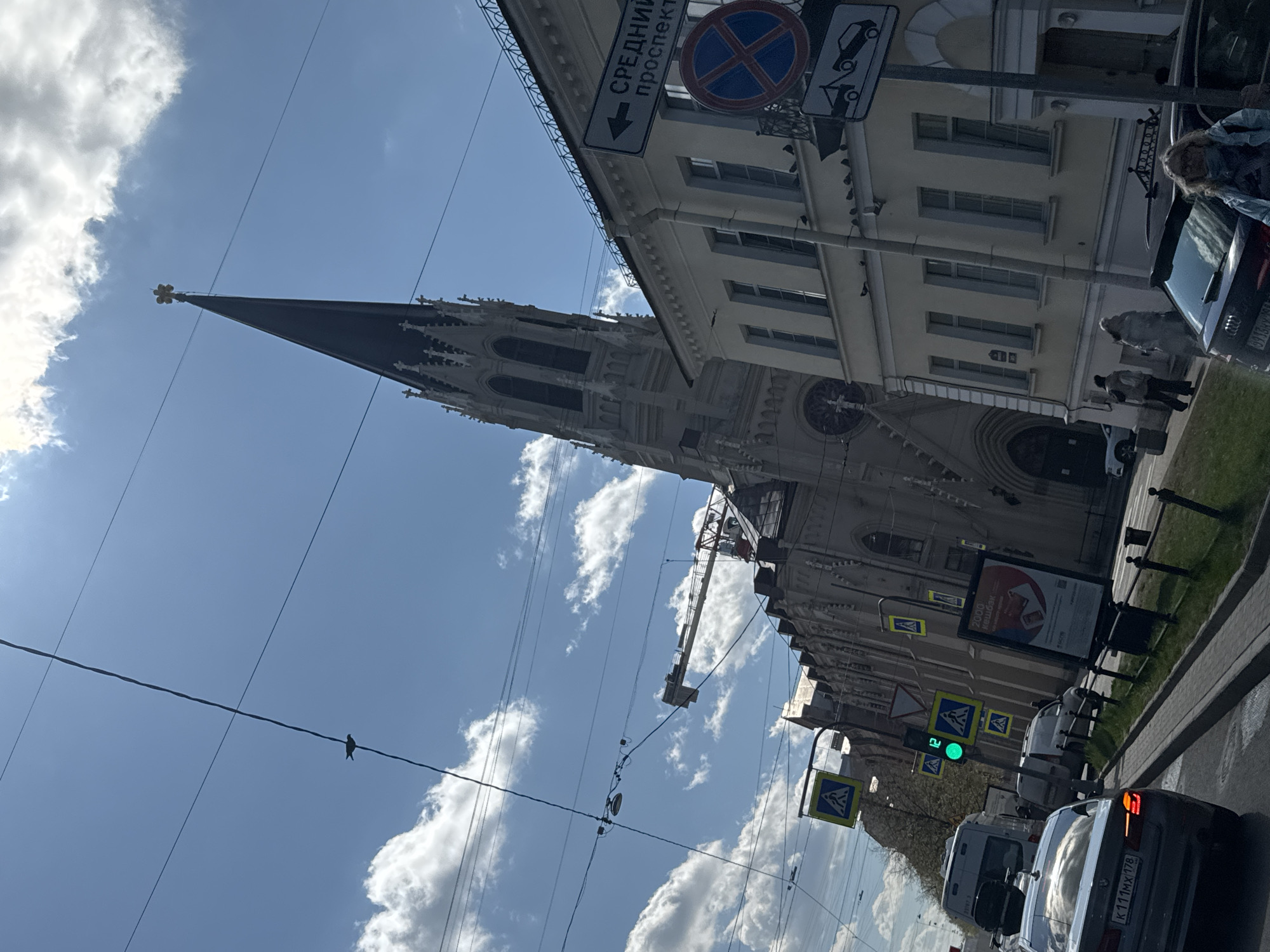The image depicts a street scene in Saint Petersburg, Russia, oriented with the top of the frame leaning to the left. The predominant subject is the Chesme Church, a Gothic Revival-style church with a tall, pointed spire visible against a backdrop of partly cloudy blue sky. Power lines crisscross the sky in the foreground.
To the right of the church is a multi-story building with a light-colored facade and multiple windows. Street signs are visible, including a 'no stopping' sign and a sign indicating 'Средний проспект' (Sredny Prospect).
On the right side of the image, there is a street with parked and moving vehicles, including cars and a white van. Pedestrians are walking along the sidewalk. Street furniture, such as lampposts and traffic signs indicating pedestrian crossings, are also present. The traffic light shows green. A bird is perched on one of the wires.