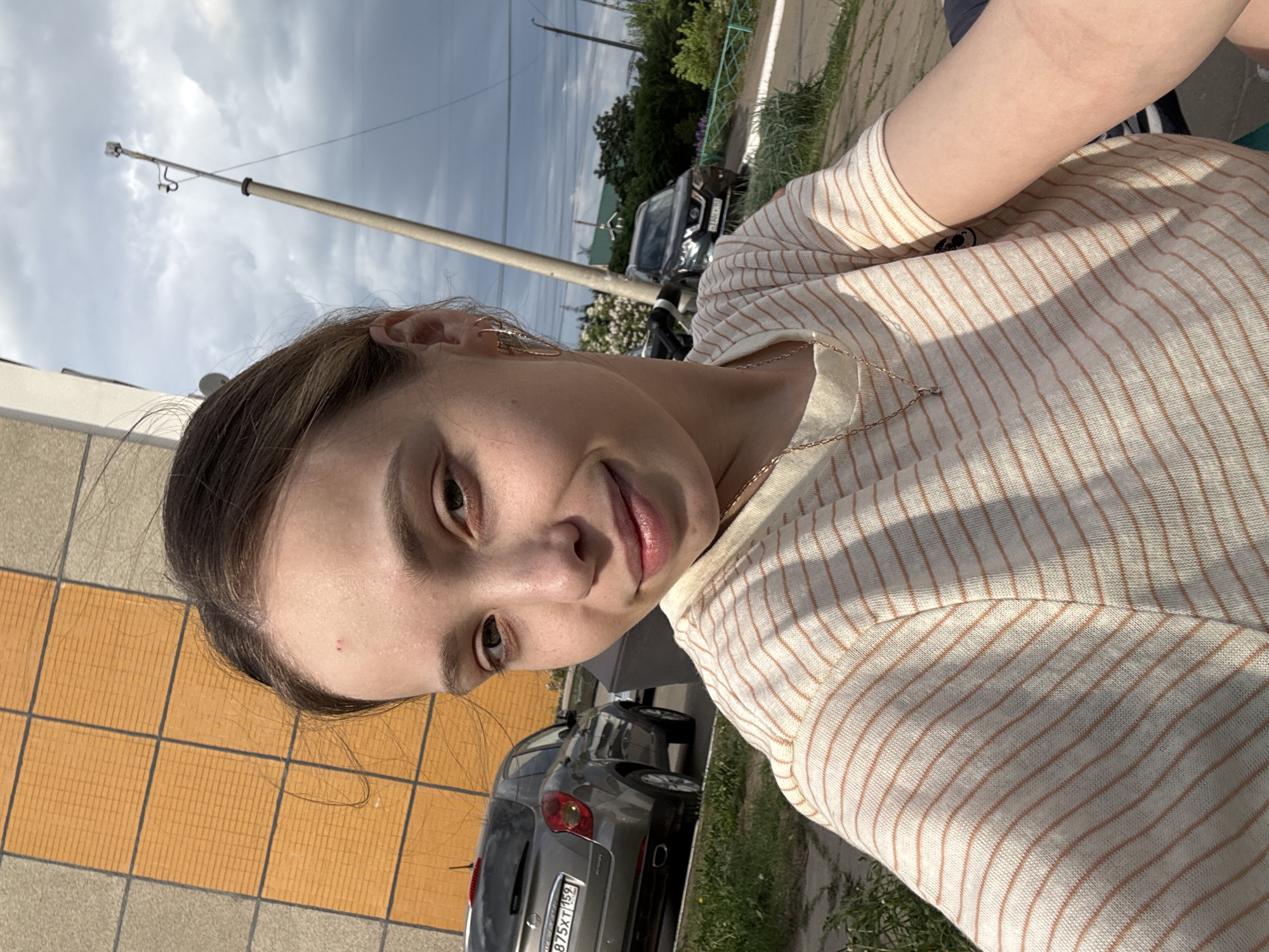 A selfie shot of a woman wearing a striped shirt and a gold necklace. The background shows a building with orange tiles, a parked gray car with the license plate number "B 875 XT 159", a green patch of grass, a road, a utility pole, and a cloudy sky. The location appears to be outdoors in the city of Chaikovsky, Russia, based on the license plate format.